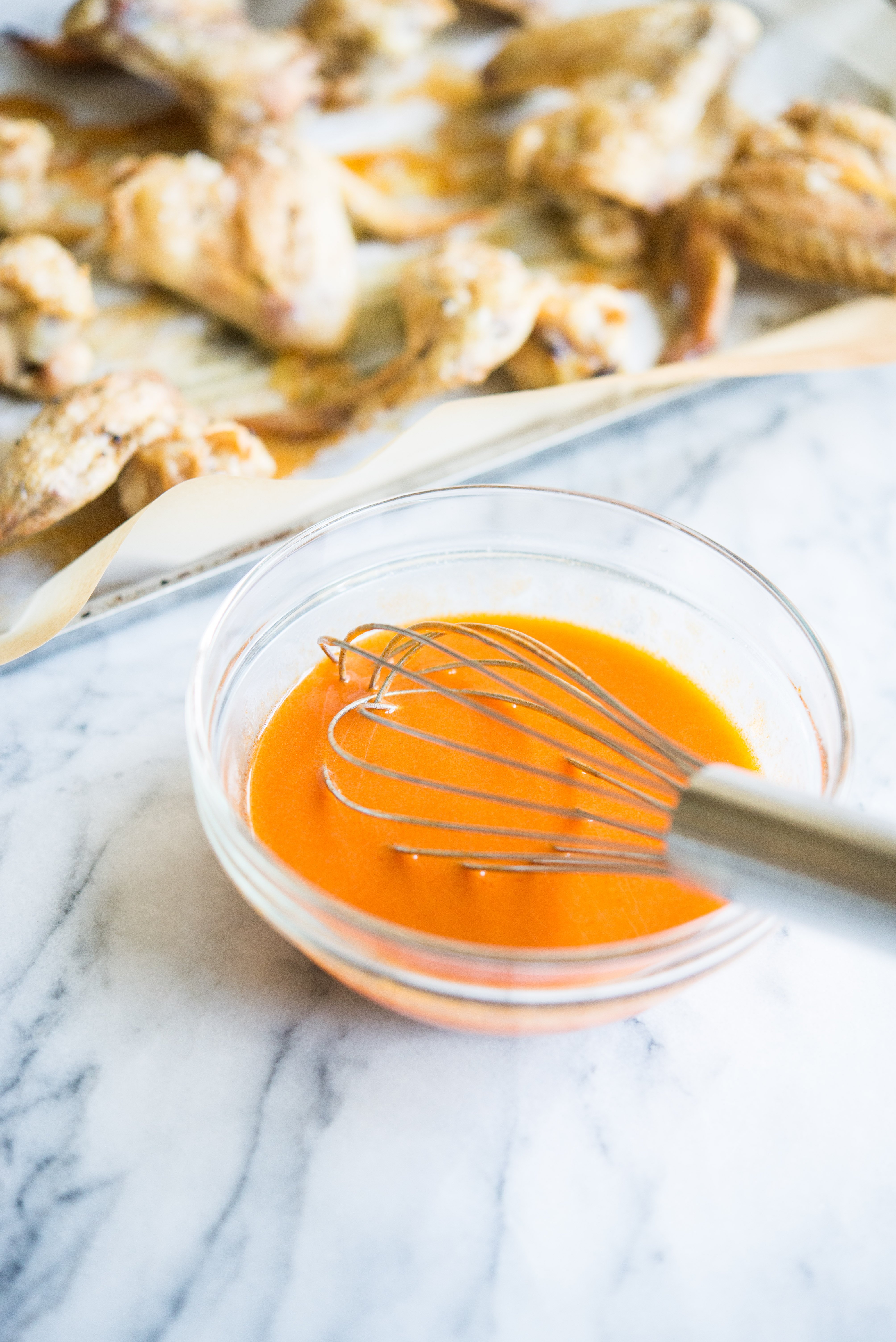 buffalo sauce in a glass bowl on a marble surface beside crispy baked chicken wings