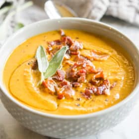 Instant pot pumpkin soup in a grey bowl topped with bacon and sage on a marble board