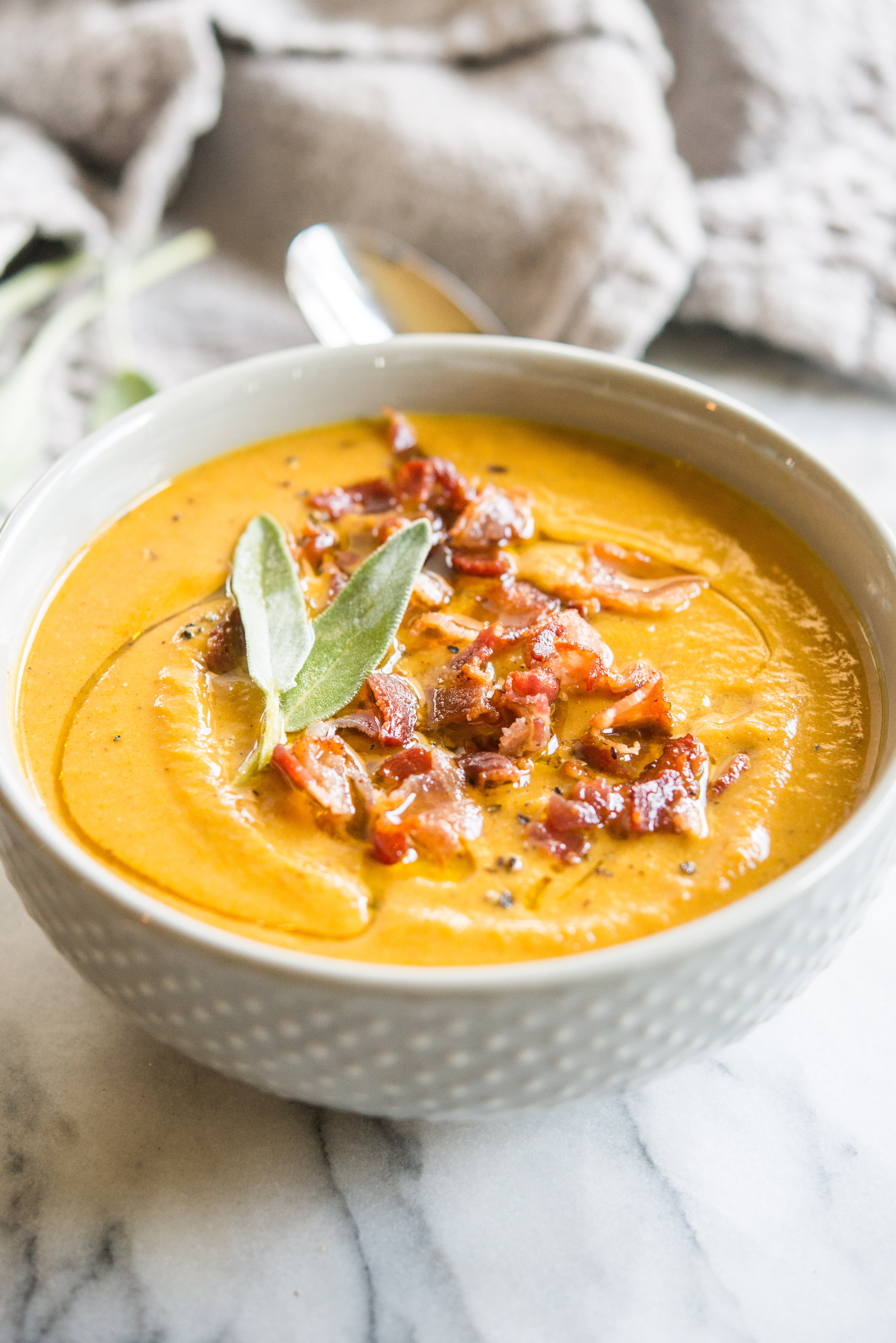 Instant pot pumpkin soup in a grey bowl topped with bacon and sage on a marble board