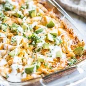 chicken enchilada casserole in a glass dish topped with diced avocados and drizzled with sour cream on a marble slab