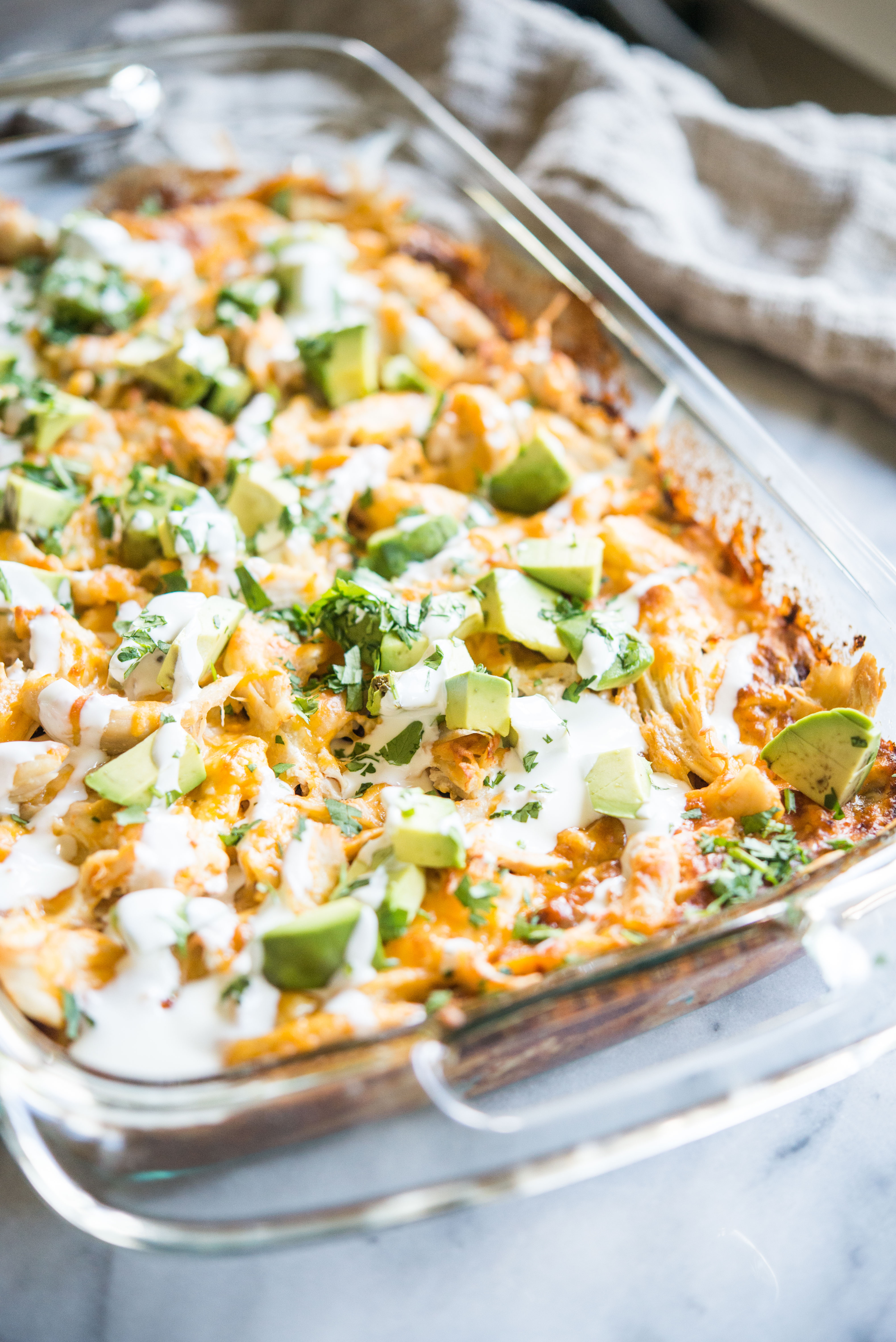 chicken enchilada casserole in a glass dish topped with diced avocados and drizzled with sour cream on a marble slab