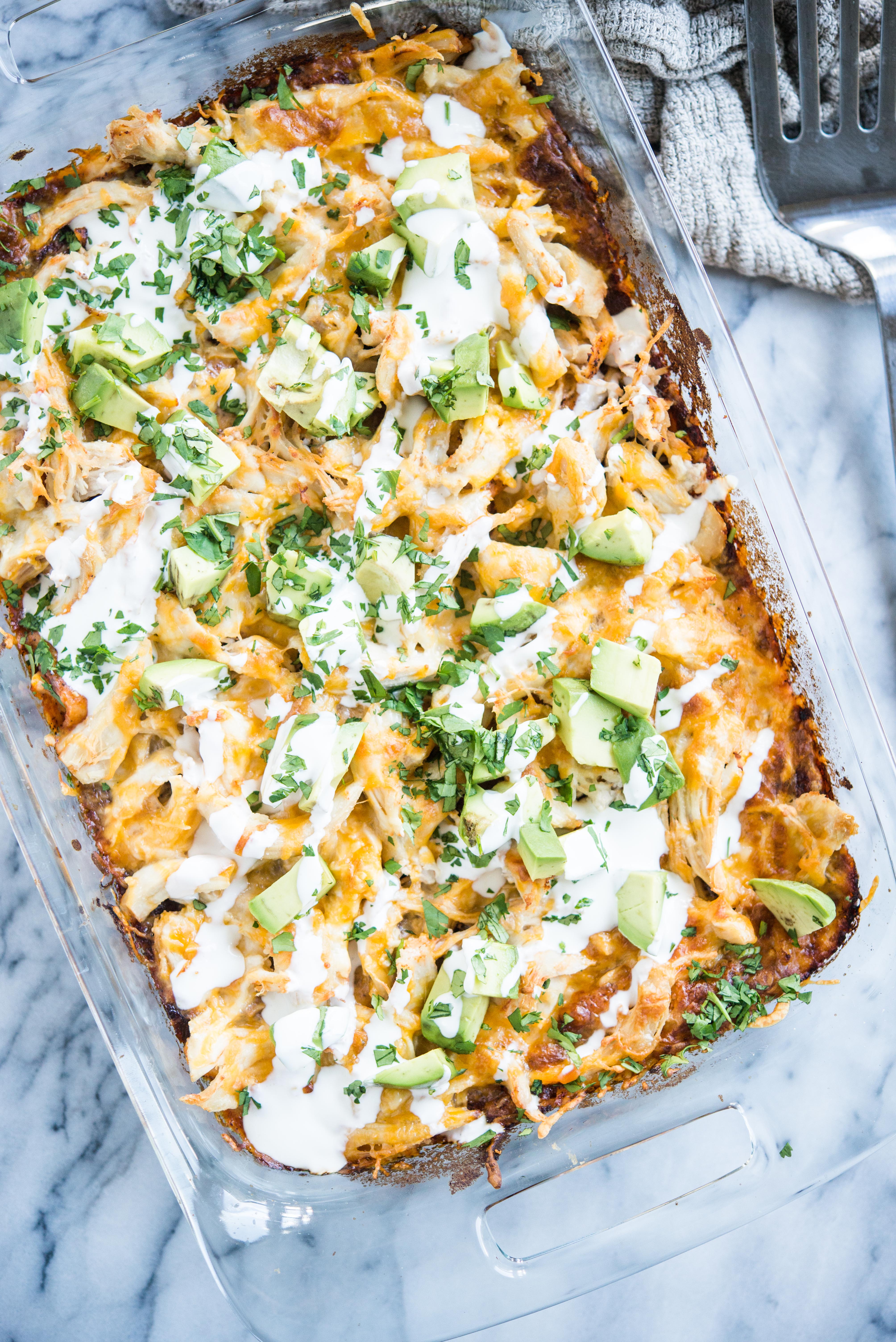 chicken enchilada casserole in a glass dish topped with diced avocados and drizzled with sour cream on a marble slab