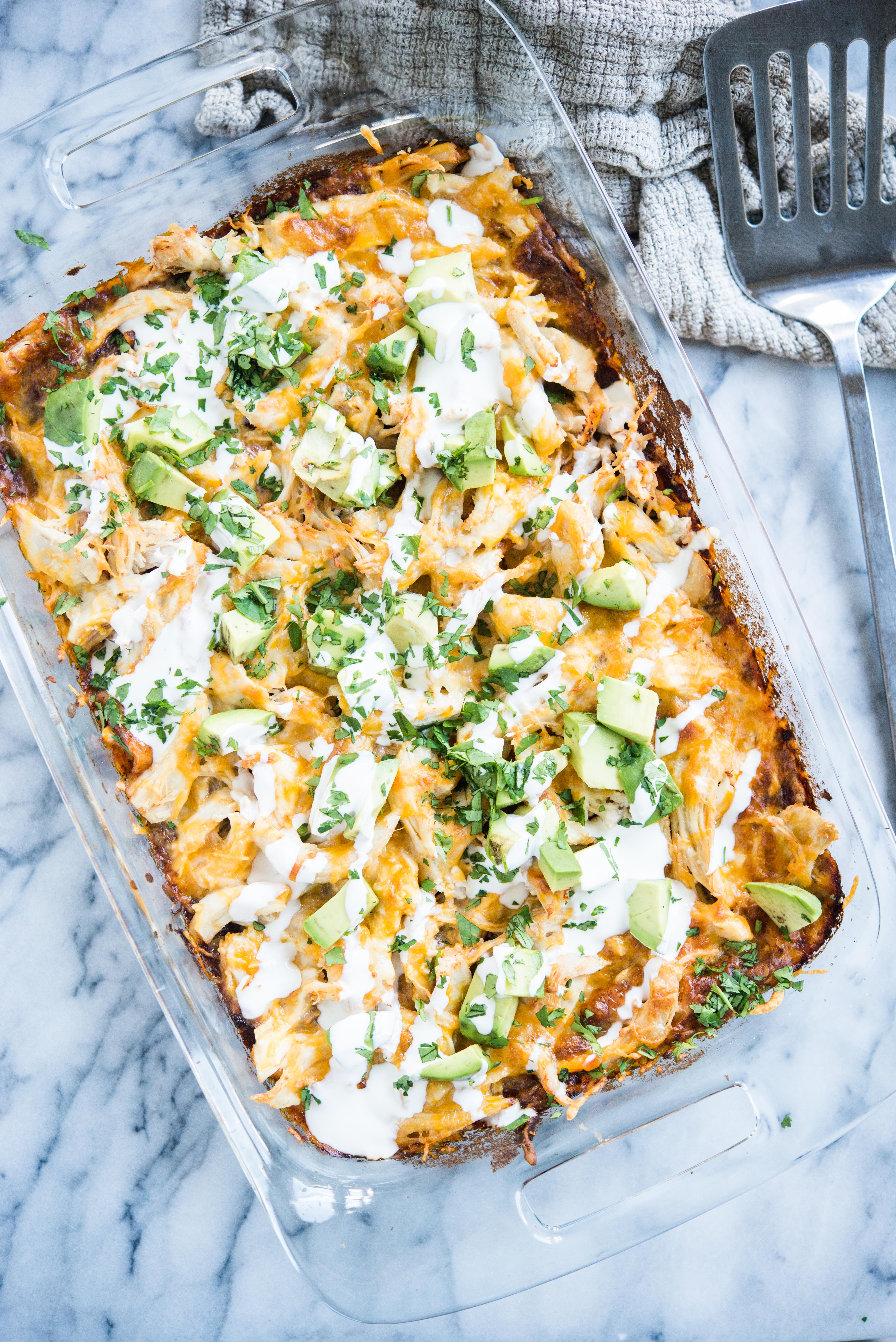 chicken enchilada casserole in a glass dish topped with diced avocados and drizzled with sour cream on a marble slab