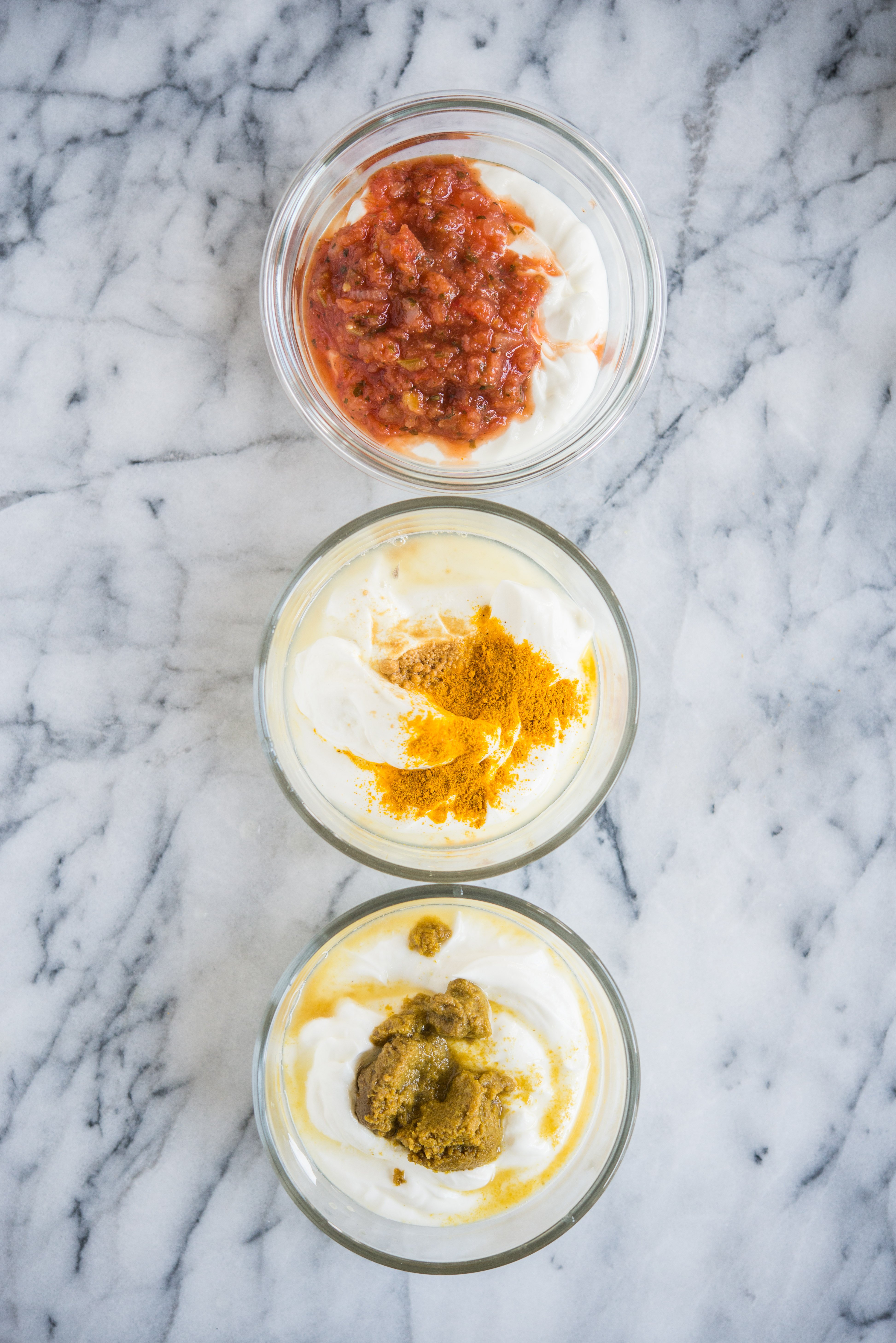three bowls of greek yogurt lined up with spices on top sitting on a marble counter