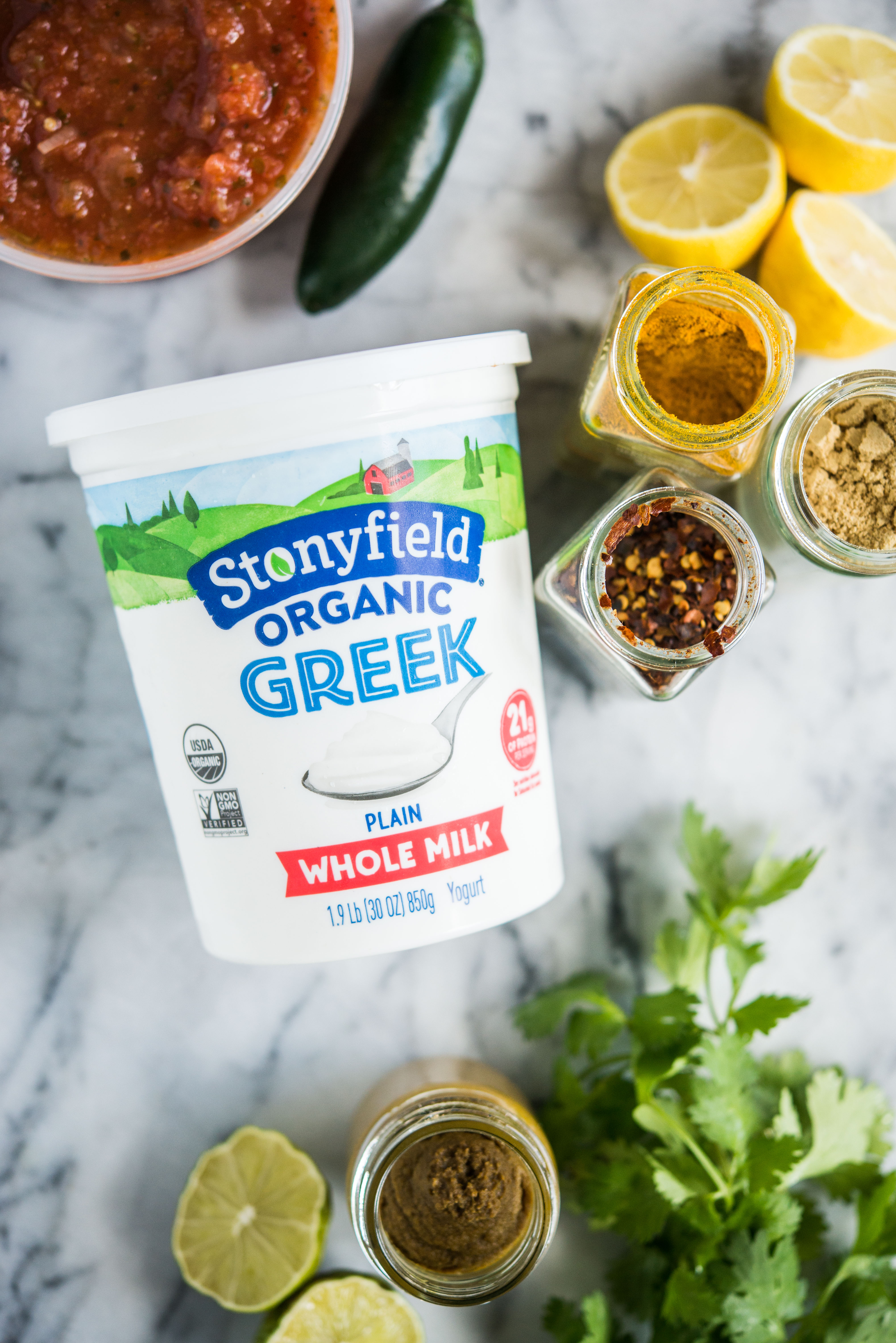 a container of stonyfield greek yogurt on a marble board surrounded by spices, lemons, limes, and salsa