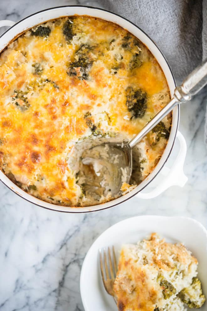 cheesy chicken broccoli rice casserole in a round white pot with a scoop taken out of it and set on a white plate on a marble surface