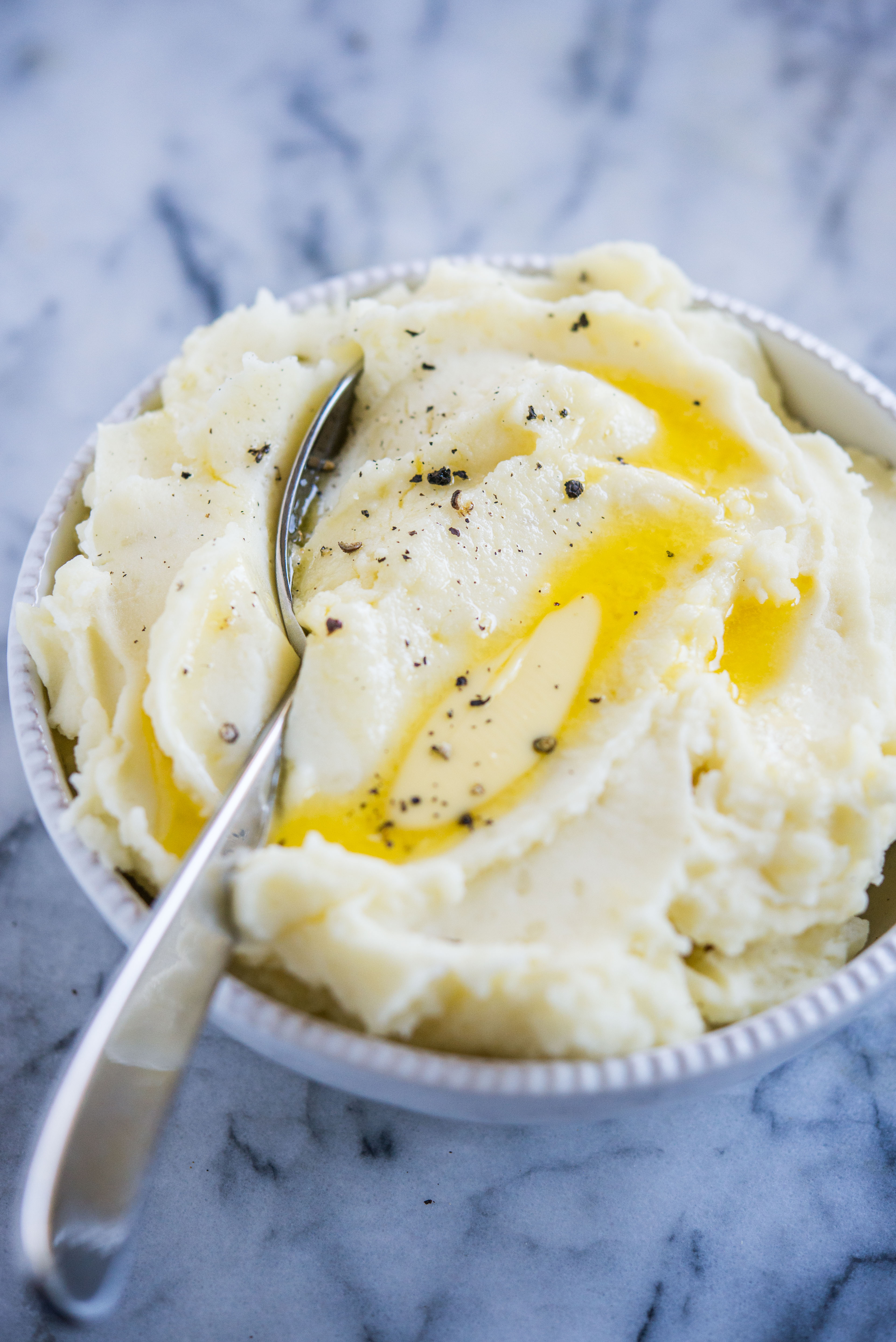 mashed potatoes in a grey bowl with pools of melted butter and a large silver serving spoon in them