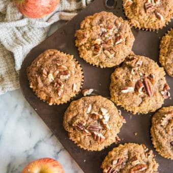 paleo apple cinnamon muffins in a muffin tin on a marble board with apples and a grey cloth surrounding them