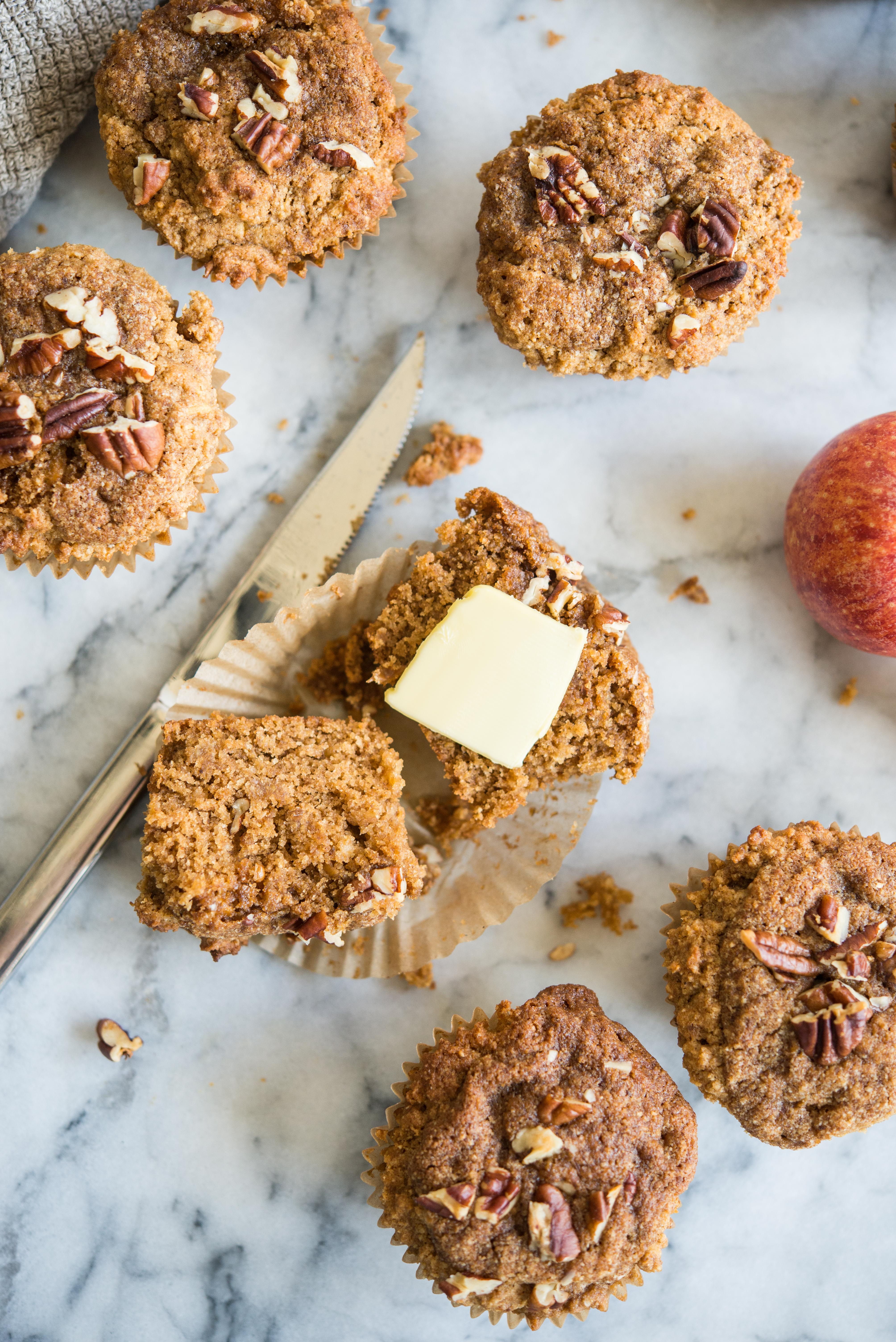 an apple cinnamon muffin cut in half with butter on top surrounded by more muffins all on a marble countertop