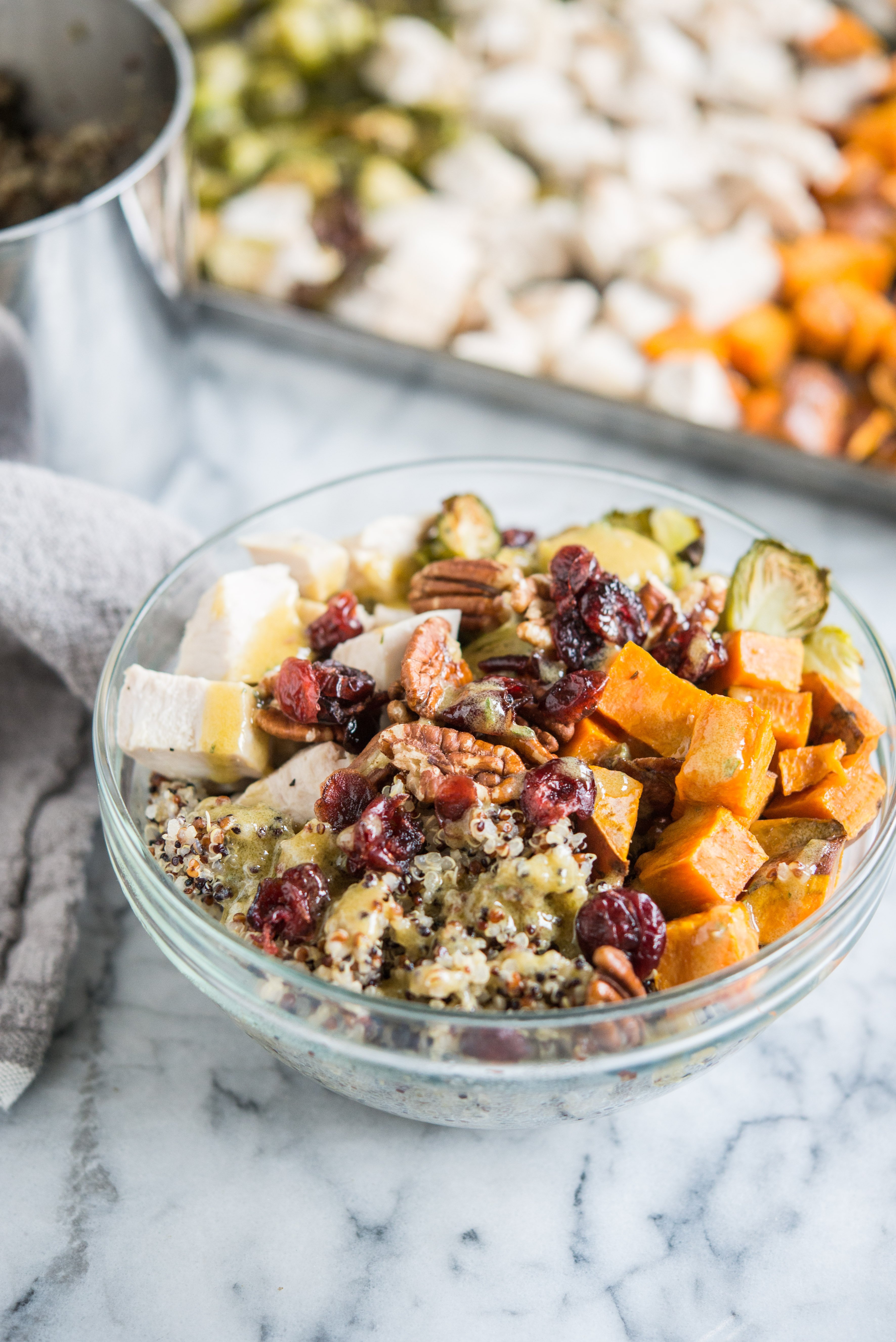 quinoa bowls with sweet potatoes, chicken, brussels spouts, pecans, and cranberries in a glass bowl on a marble countertop in front of a sheet pan with cubed chicken and sweet potatoes