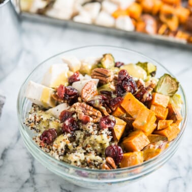 quinoa bowls with sweet potatoes, chicken, brussels spouts, pecans, and cranberries in a glass bowl on a marble countertop in front of a sheet pan with cubed chicken and sweet potatoes