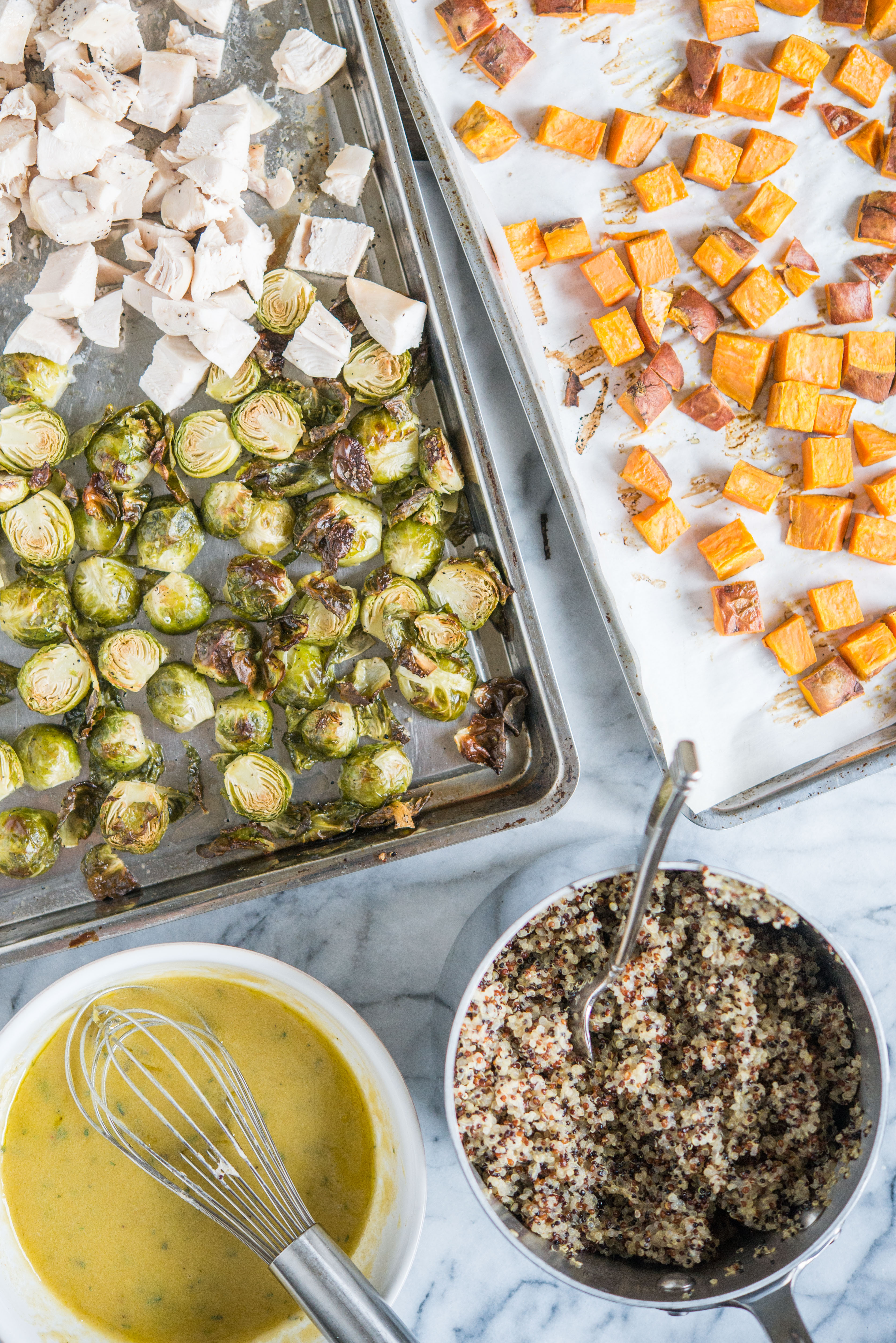 cubed chicken breast, roasted brussels sprouts, and roasted sweet potatoes on two sheet pans next to a pot of quinoa and a bowl full of dijon dressing