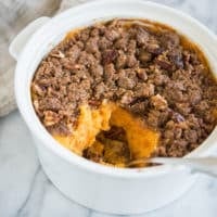 sweet potato casserole with pecan topping in a white casserole dish with a scoop taken out of it sitting on a marble surface
