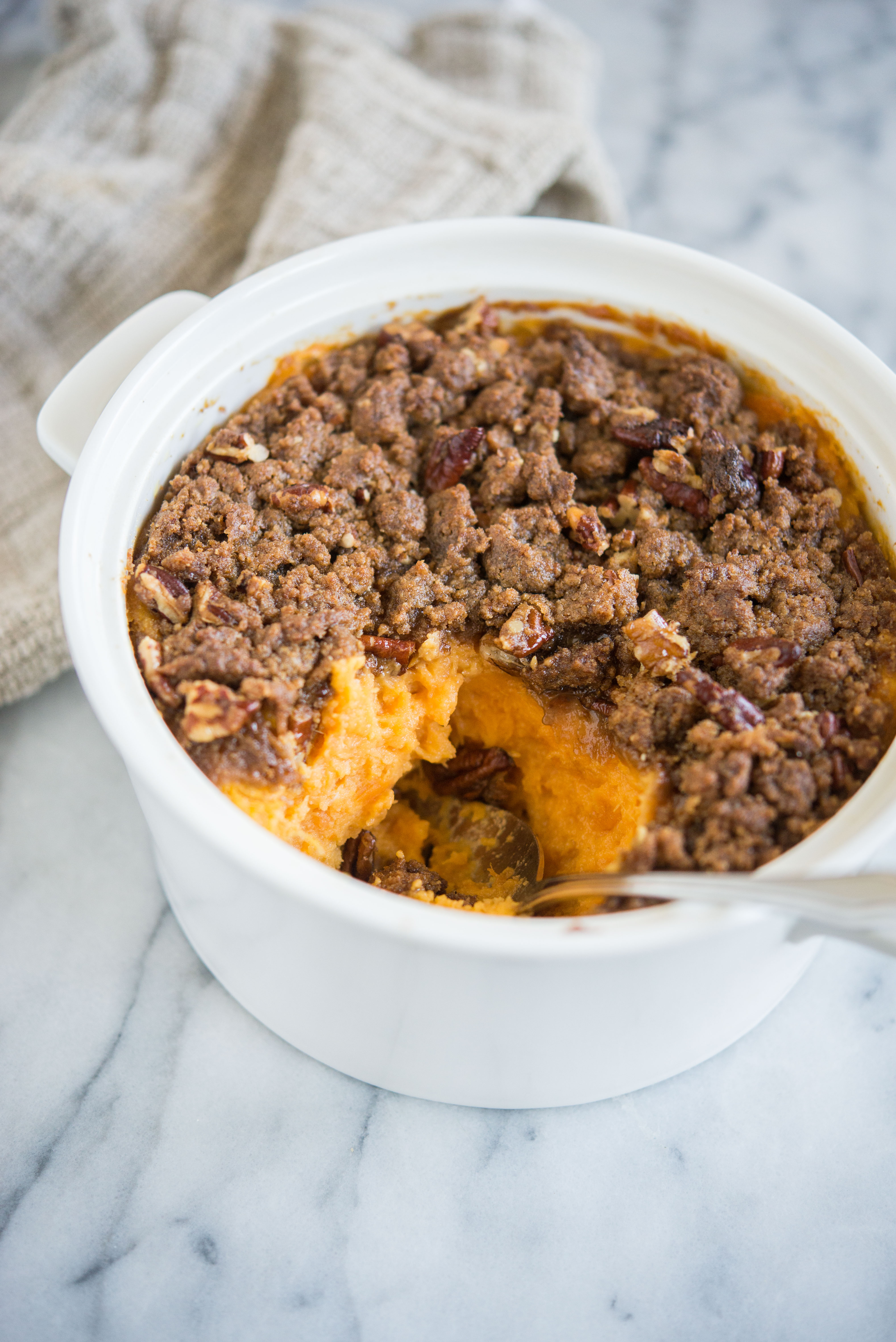 sweet potato casserole with pecan topping in a white casserole dish with a scoop taken out of it sitting on a marble surface
