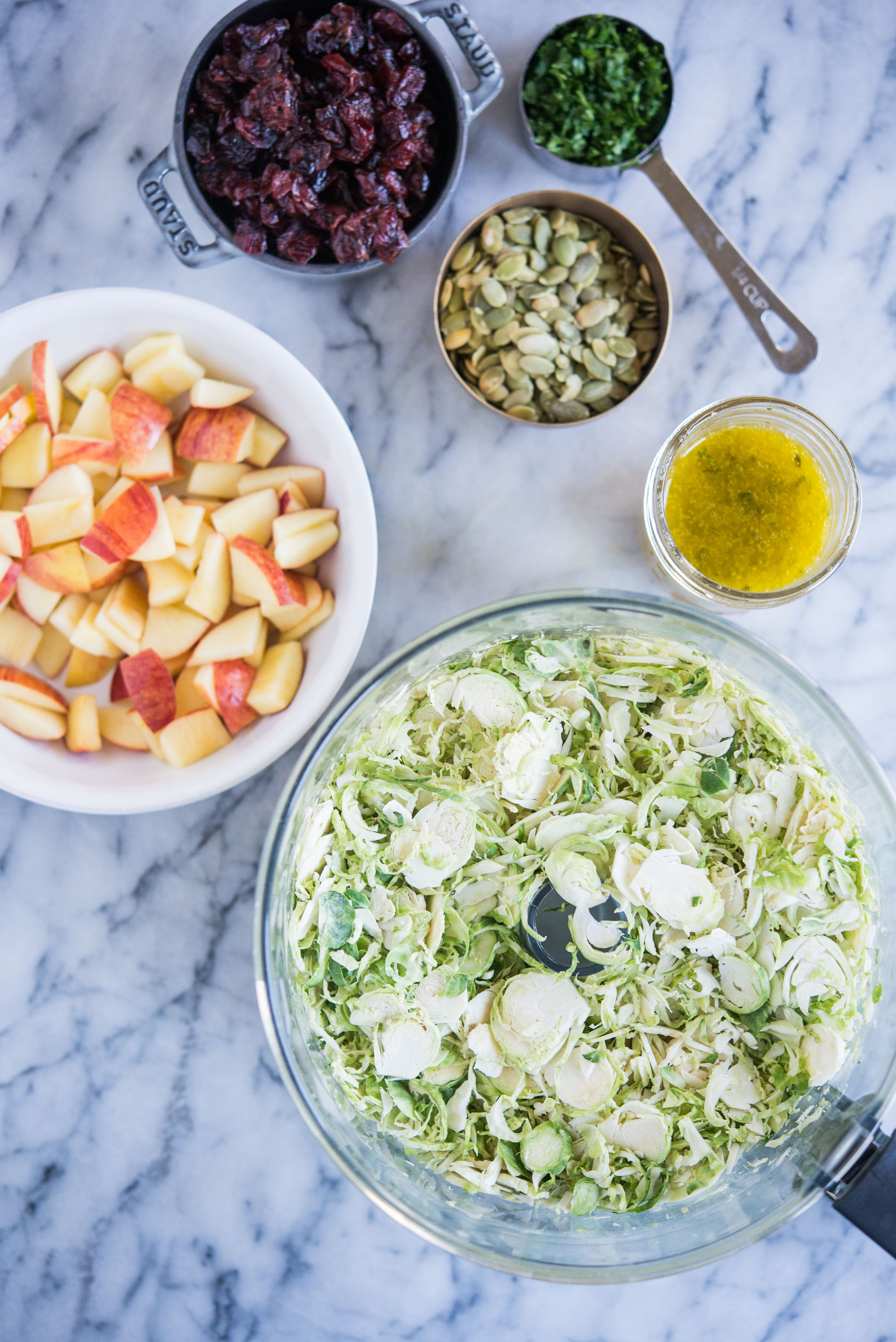 ingredients for shredded brussels sprouts salad - shredded brussels sprouts, diced apples, pumpkin seeds, dried cranberries, parsley, and apple cider vinaigrette in separate bowls on a marble surface