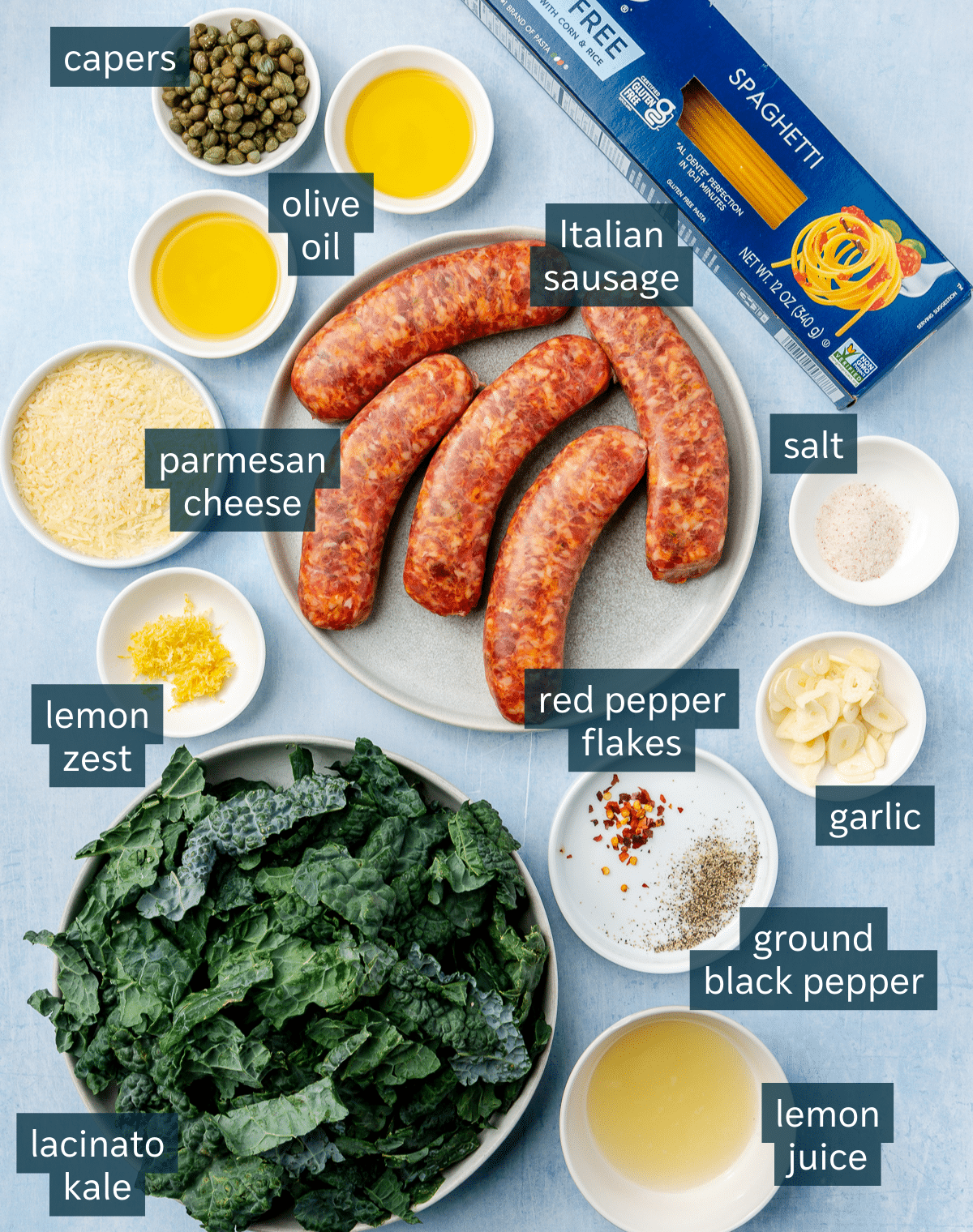 Ingredients for pasta with sausage and kale sit on a variety of plates and in a variety of bowls. A box of spaghetti sits close to the top.