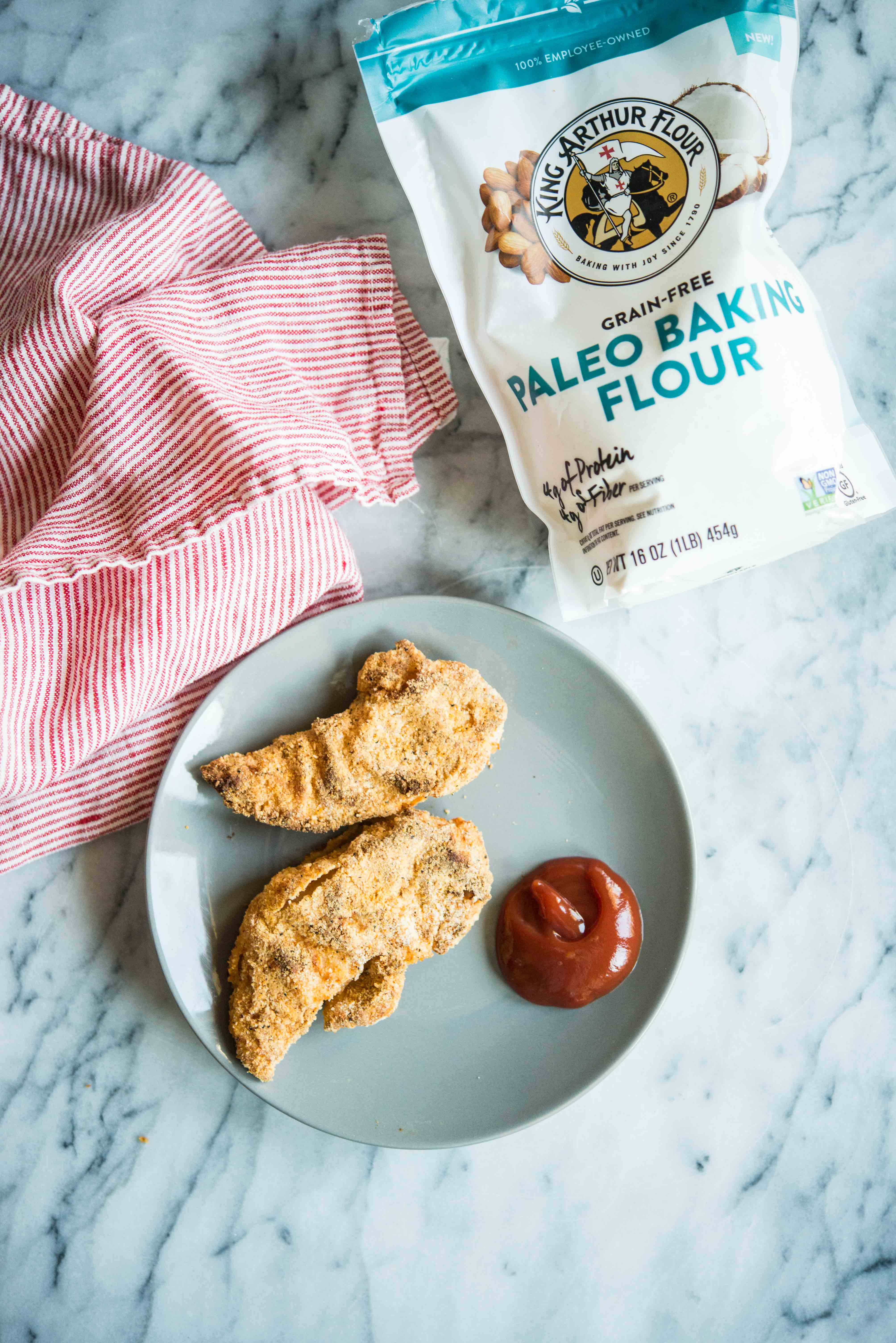 two chicken tenders on a grey plate with ketchup on a marble surface with a bag of paleo baking flour beside it and a red striped cloth