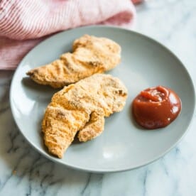 two paleo chicken tenders on a plate with ketchup on a marble surface with a red striped towel in the background