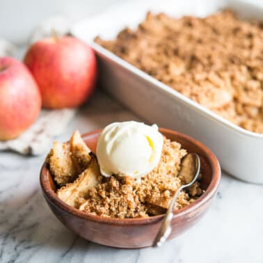 paleo apple crisp in a wooden bowl topped with a scoop of ice cream on a marble surface