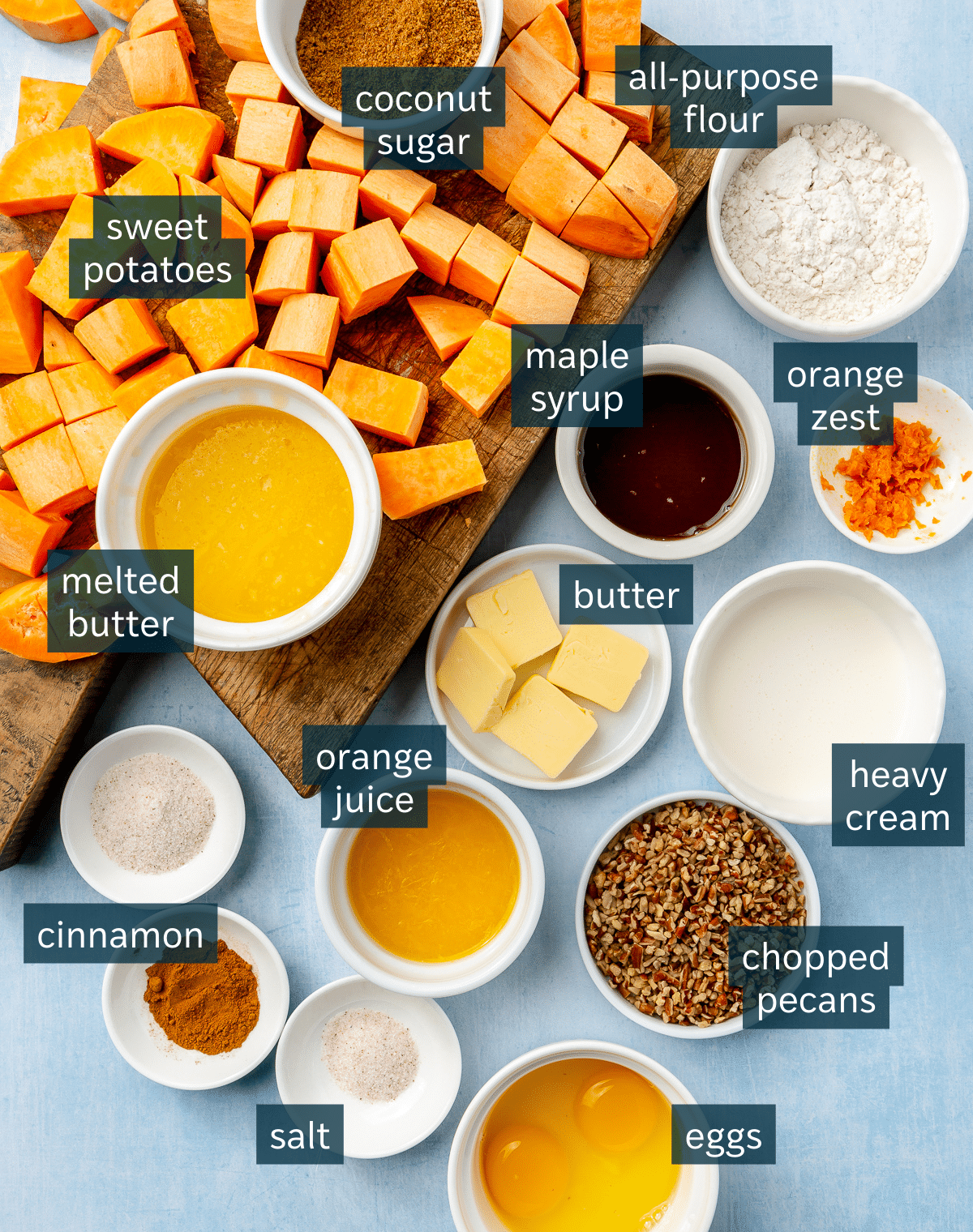 Ingredients for sweet potato casserole sit in a variety of bowls. Cubed sweet potato lays on a brown chopping block.