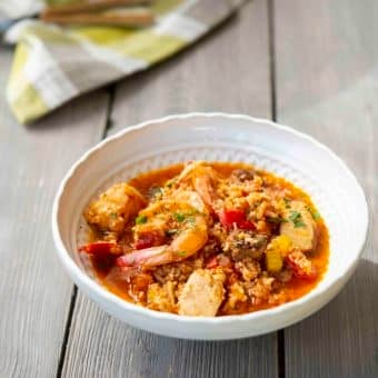 shrimp jambalaya in a white bowl with fresh chopped parsley on top on a wooden table with a green and white checked towel behind it