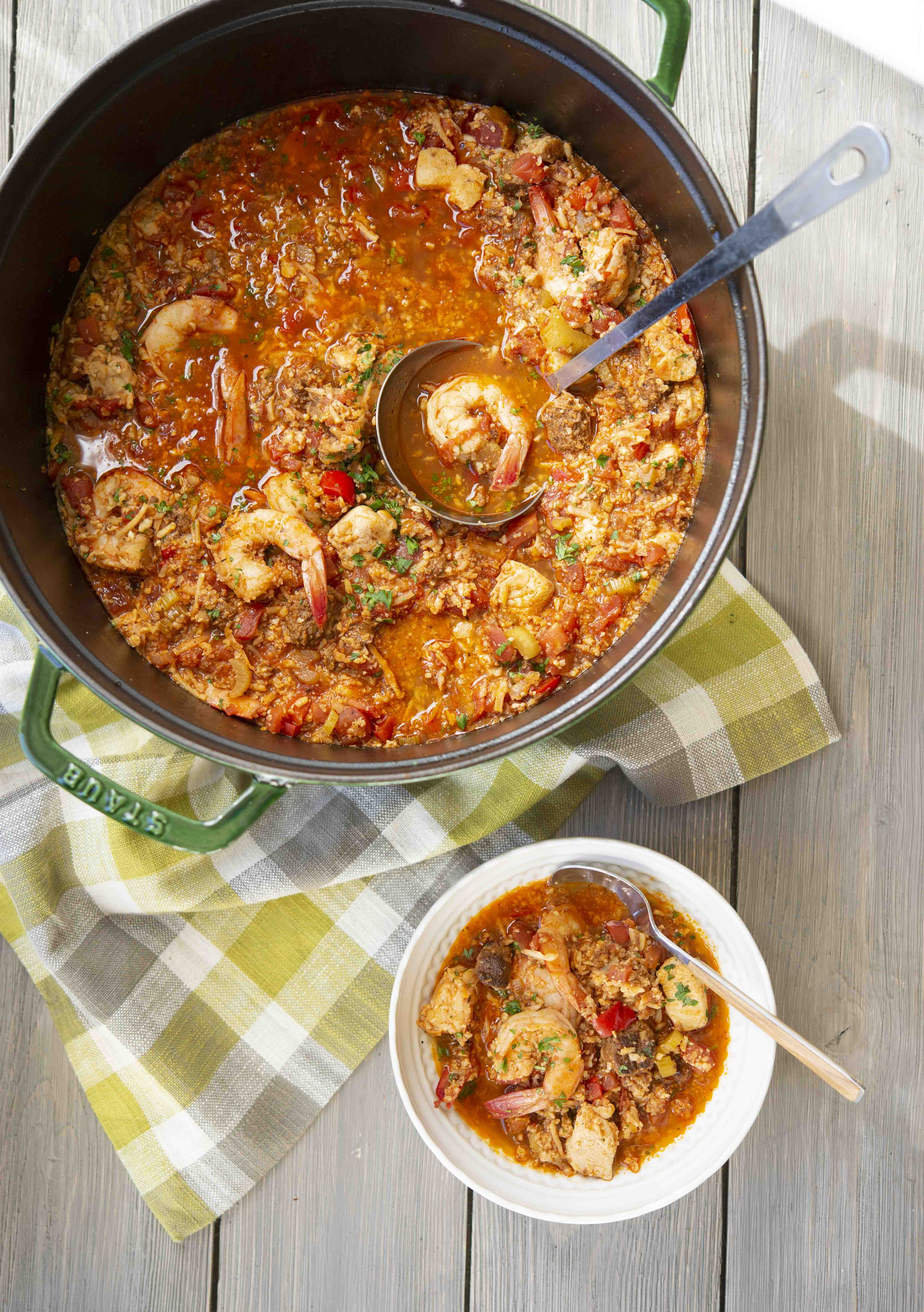 cajun shrimp jambalaya in a large green pot next to a white bowl filled with jambalaya on a wooden table with a green and grey checked towel