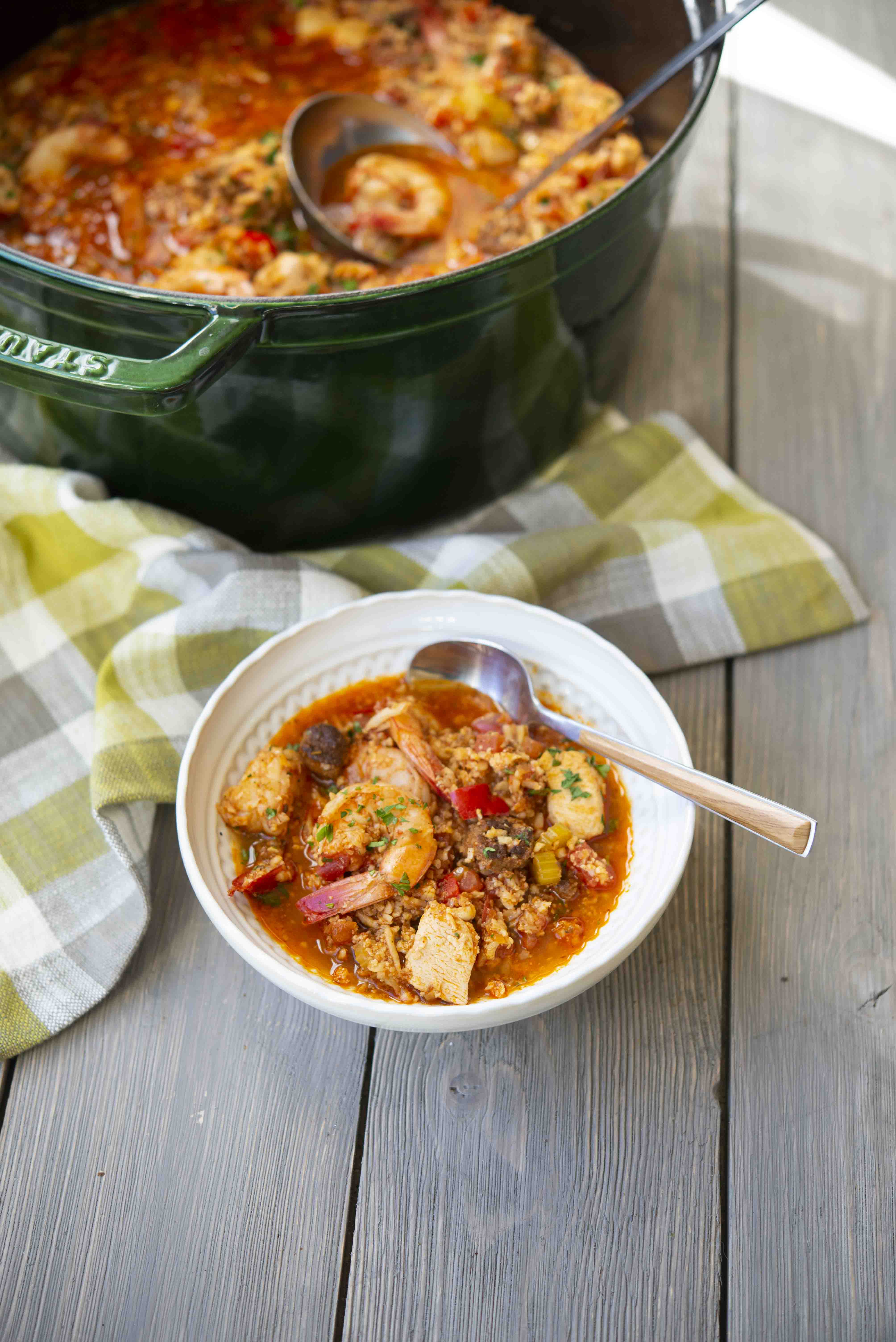 cajun shrimp jambalaya in a large green pot next to a white bowl filled with jambalaya on a wooden table with a green and grey checked towel