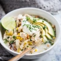 white chicken chili in a white bowl topped with a scoop of sour cream, chopped cilantro, avocado slices, and a halved lime, on a marble surface with a grey towel behind it