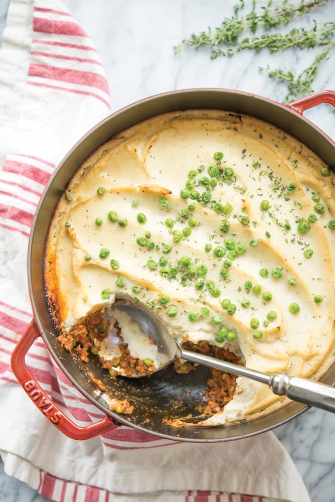 cottage pie with cauliflower mash in a red cast iron pot on a white background with a red and white striped towel lying next to it