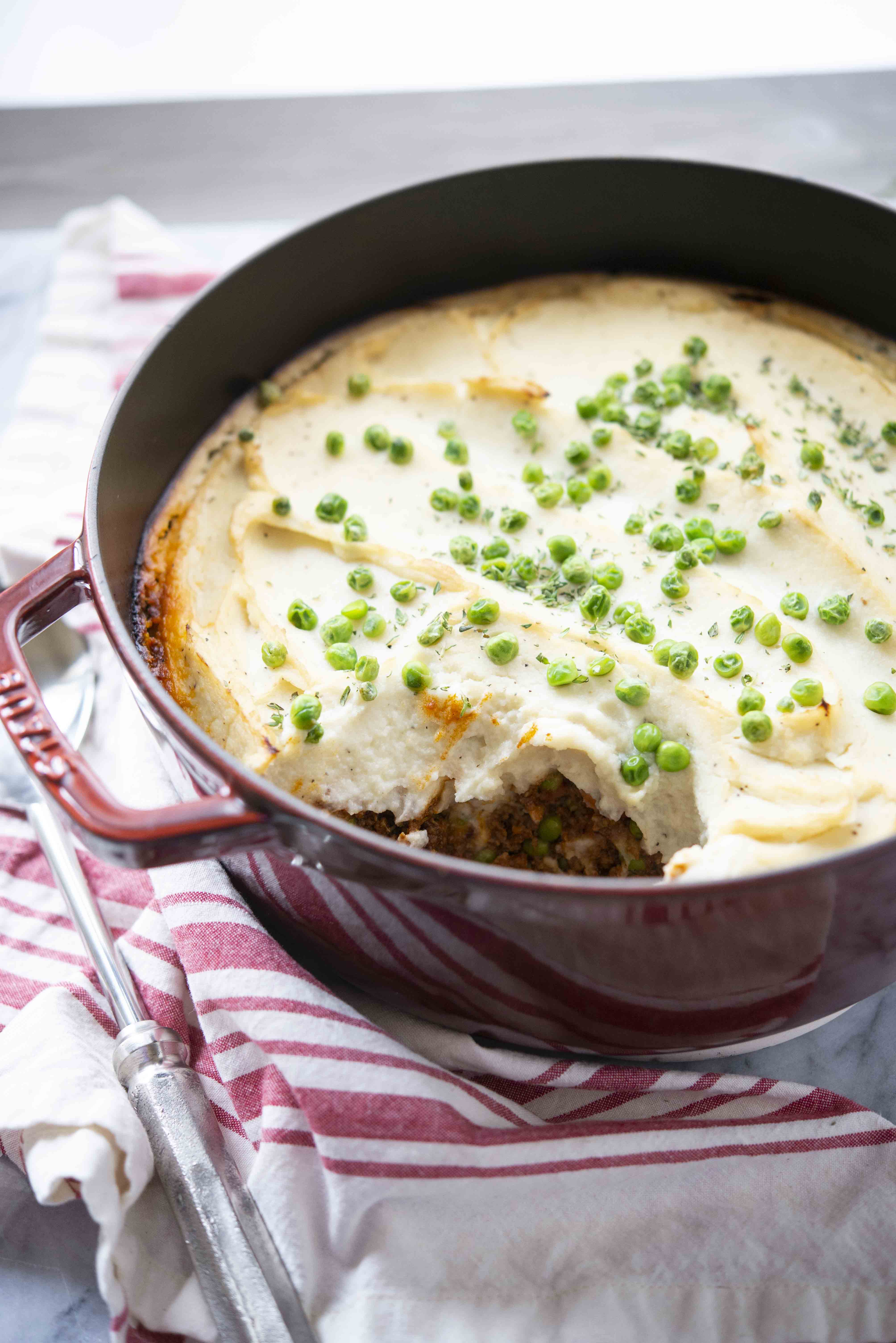 cottage pie with cauliflower mash in a red cast iron pot on a white background with a red and white striped towel lying next to it