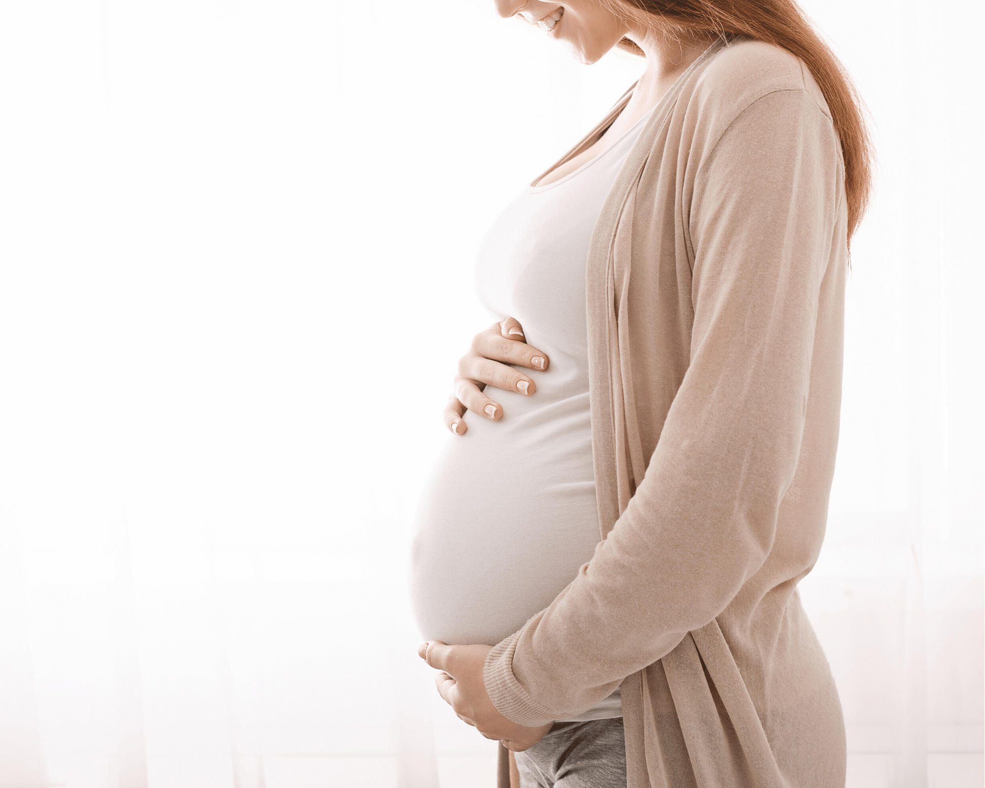 pregnant woman in a white tank top and tan cardigan holding her belly