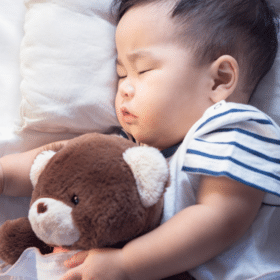 toddler in a striped shirt on a bed with a white pillow and sheets sleeping and cuddling a teddy bear - baby sleep