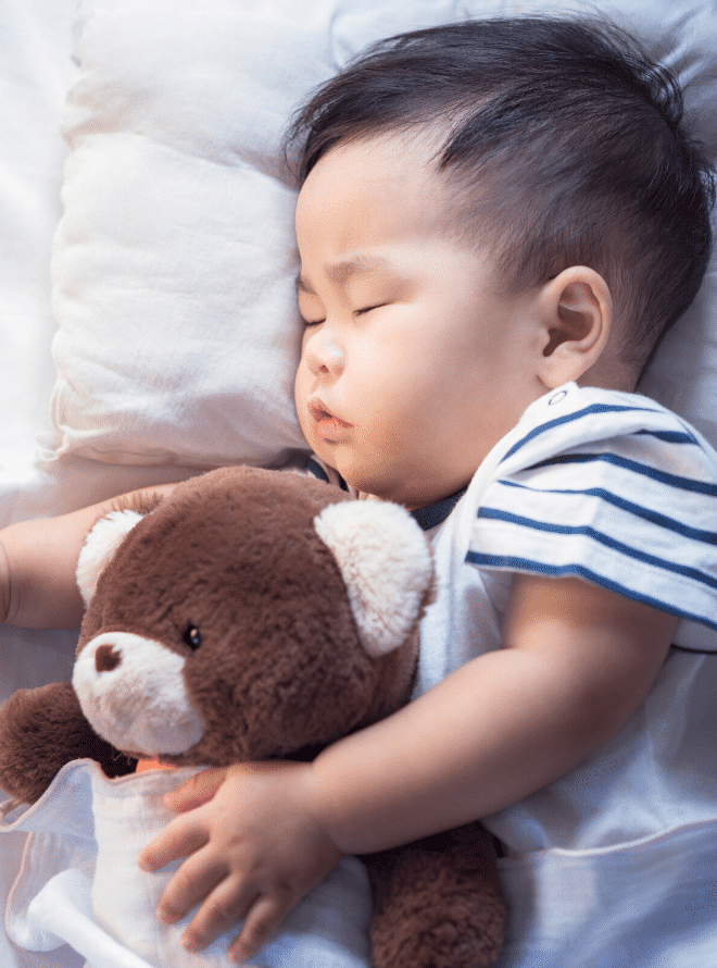 toddler in a striped shirt on a bed with a white pillow and sheets sleeping and cuddling a teddy bear - baby sleep