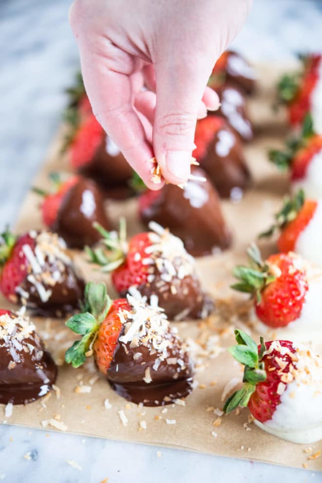 hand sprinkling toppings on top of chocolate covered strawberries sitting on parchment paper on a marble surface