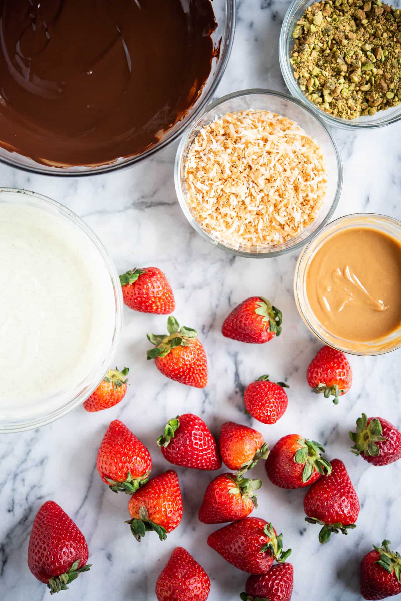 dark chocolate, white chocolate, pistachios, toasted coconut, and peanut butter in glass bowls next to strawberries on a marble surface