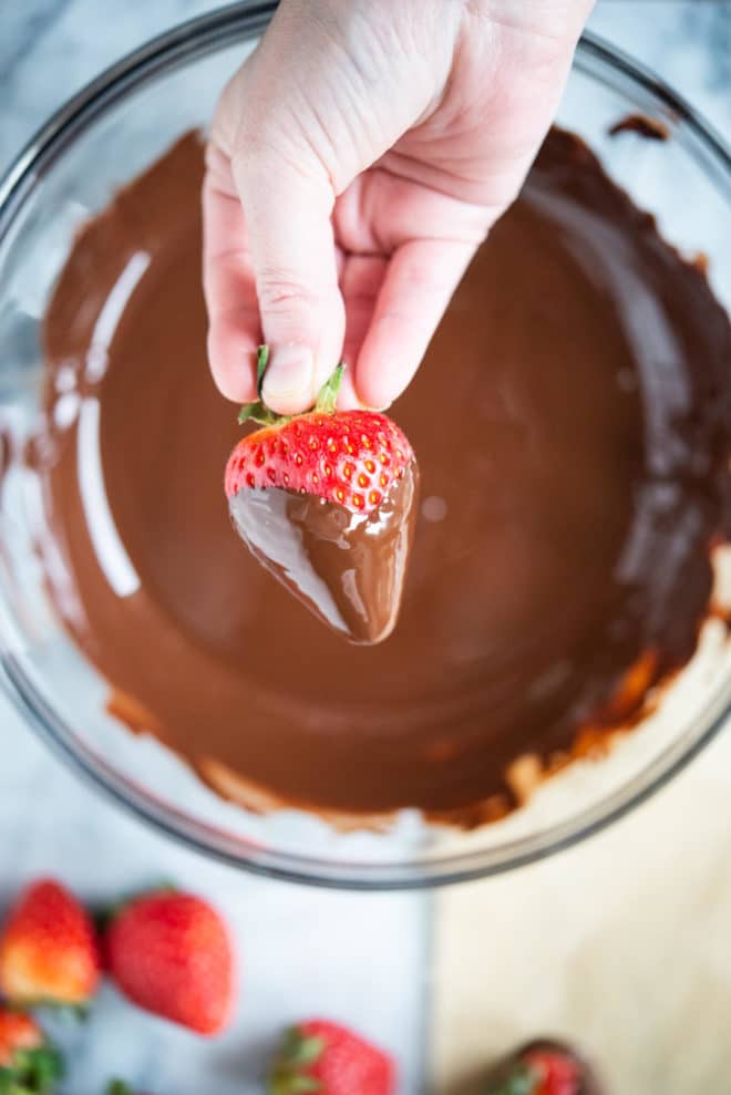 how to make chocolate covered strawberries - hand holding a chocolate dipped strawberry over a glass bowl filled with melted chocolate
