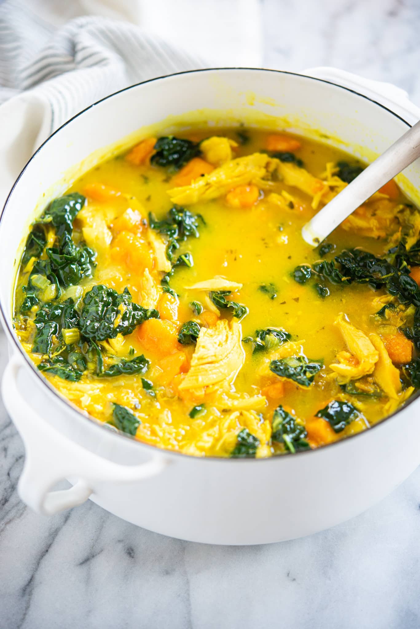 healing chicken soup with yellow turmeric broth, sweet potatoes, and kale in a white pot on a marble surface