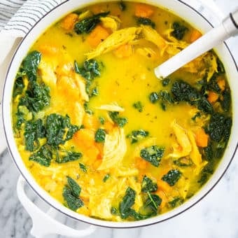 healing chicken soup with yellow turmeric broth, sweet potatoes, and kale in a white pot on a marble surface