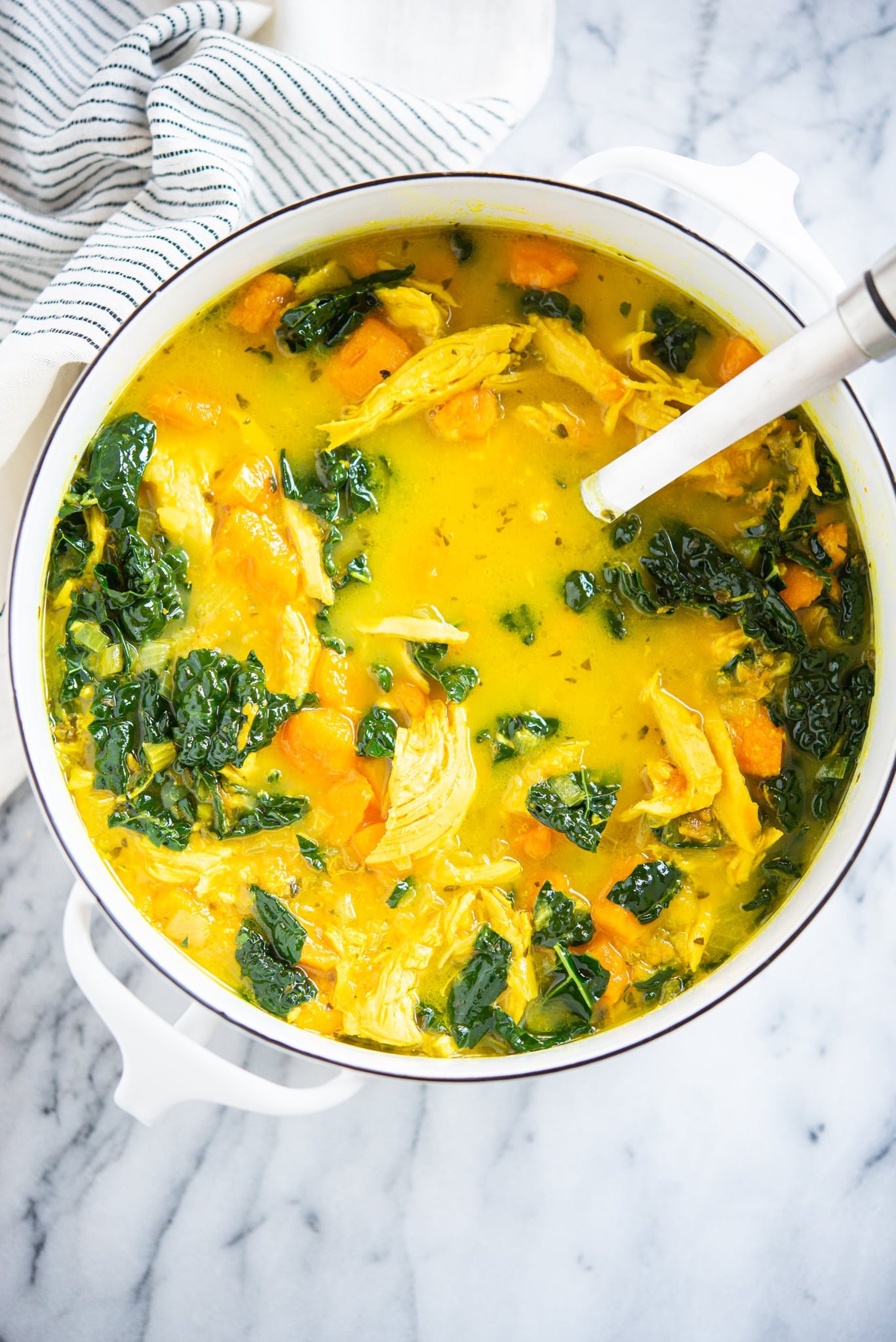 healing chicken soup with yellow turmeric broth, sweet potatoes, and kale in a white pot on a marble surface