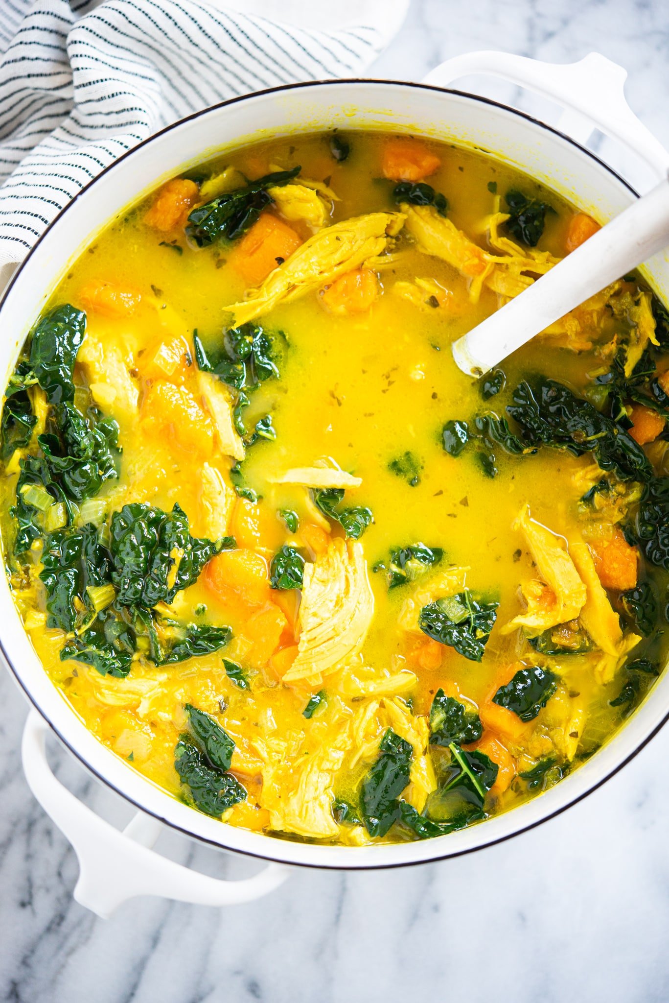 healing chicken soup with yellow turmeric broth, sweet potatoes, and kale in a white pot on a marble surface