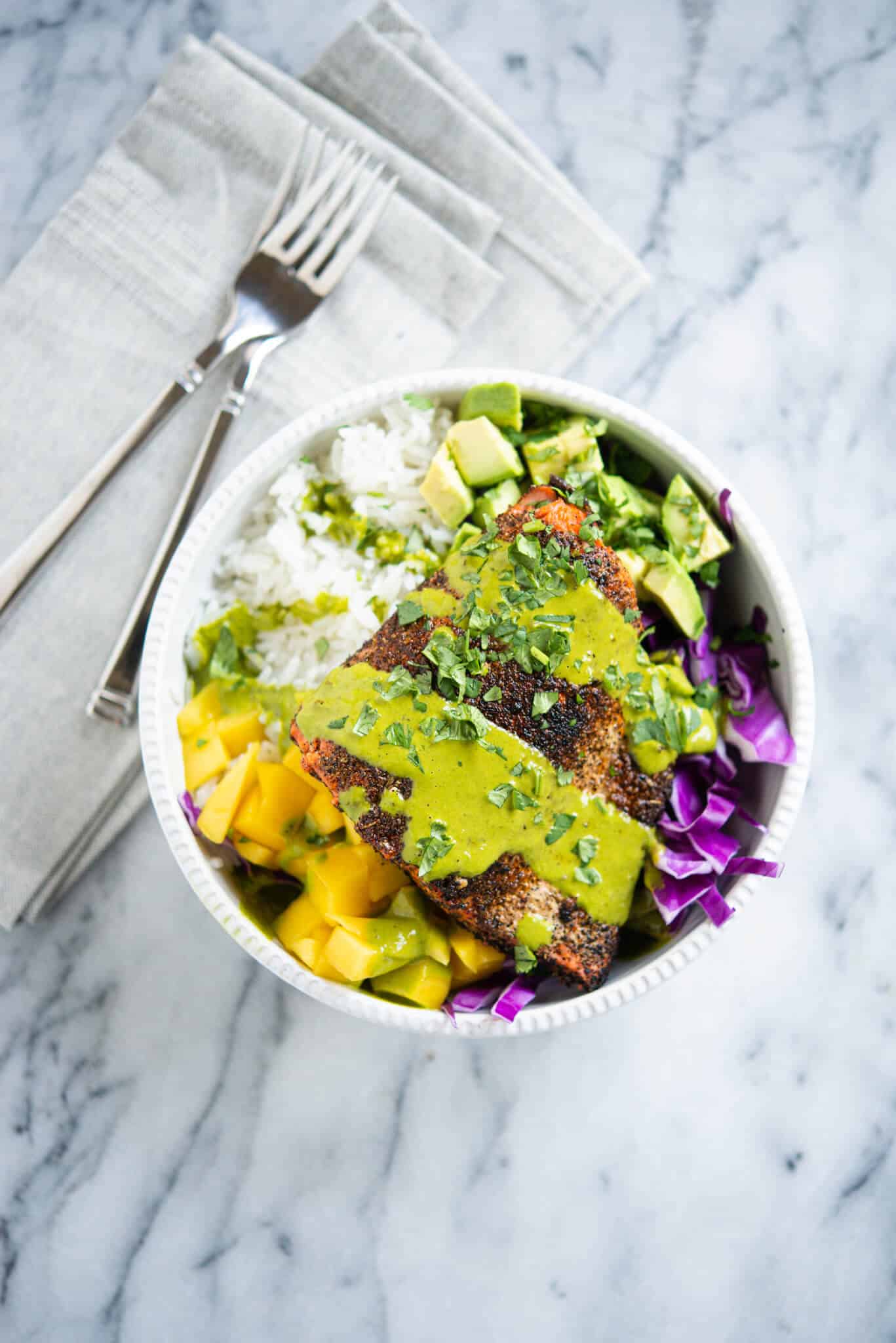 blackened salmon in a white bowl with rice, purple cabbage, mango, avocado and a green sauce sitting on a marble surface