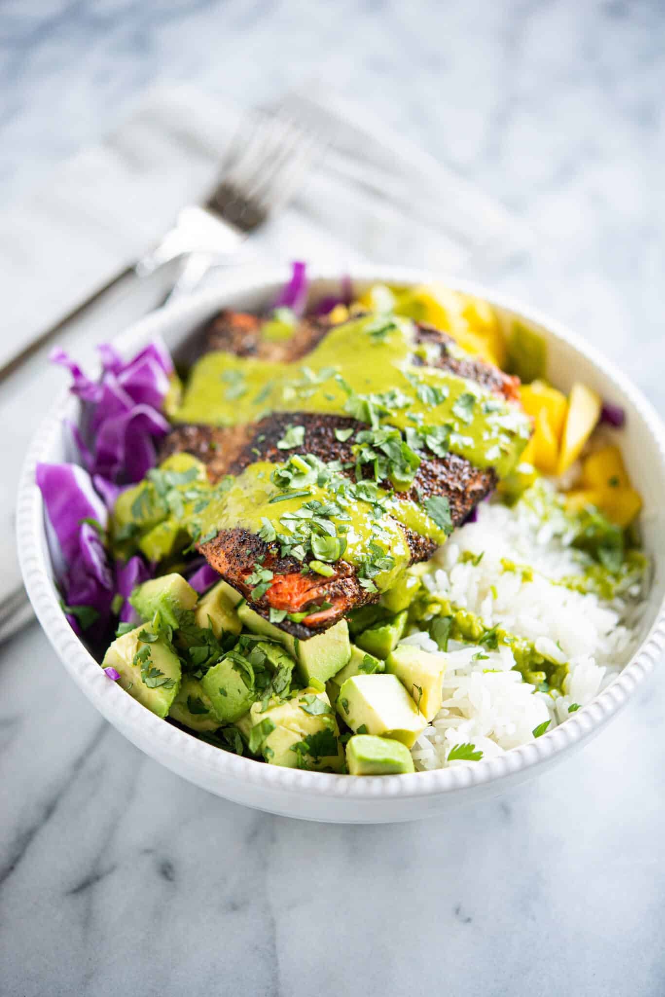 blackened salmon in a white bowl with rice, purple cabbage, mango, avocado and a green sauce sitting on a marble surface