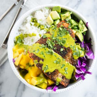 blackened salmon in a white bowl with rice, purple cabbage, mango, avocado and a green sauce sitting on a marble surface