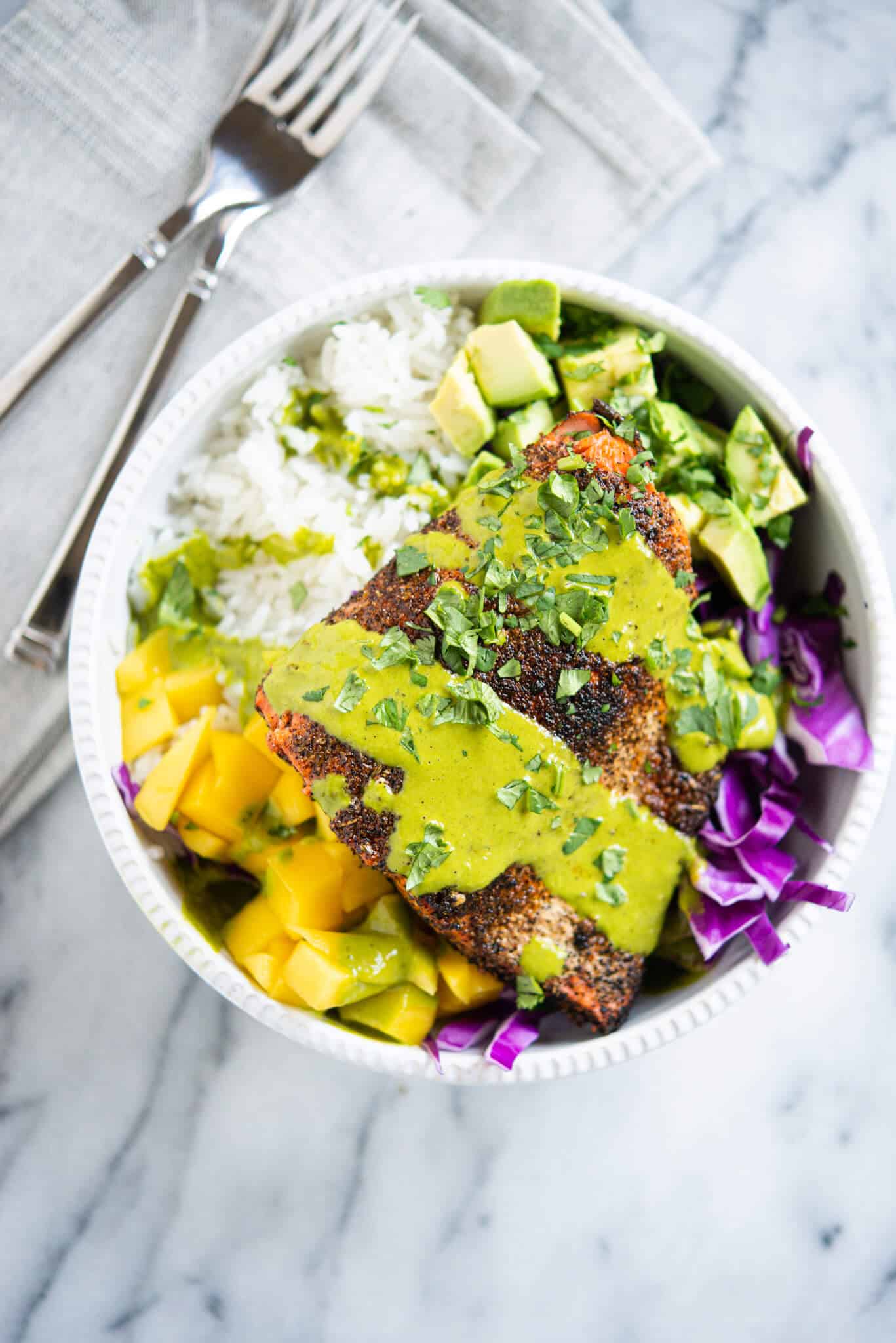 blackened salmon in a white bowl with rice, purple cabbage, mango, avocado and a green sauce sitting on a marble surface