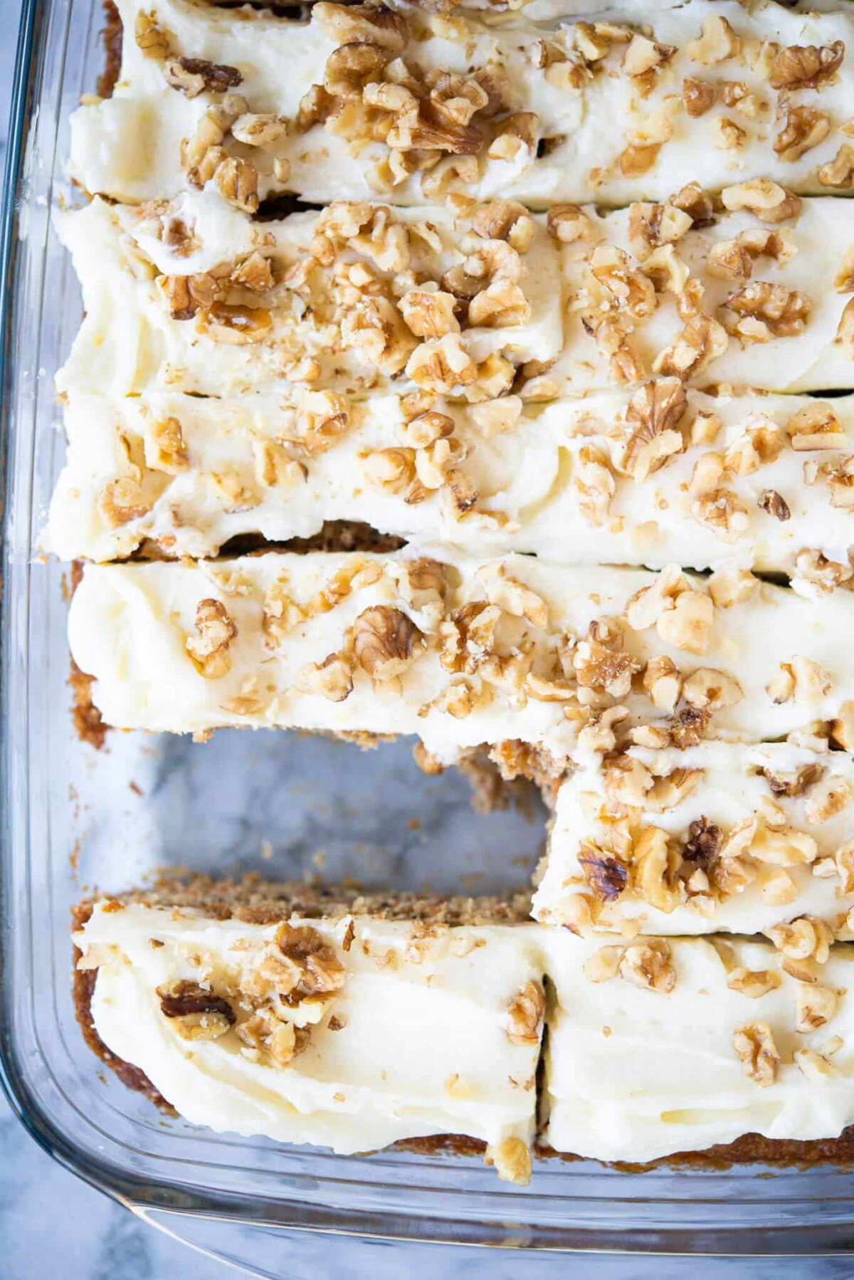 iced carrot cake bars cut into pieces with one piece missing in a clear baking dish on a marble surface