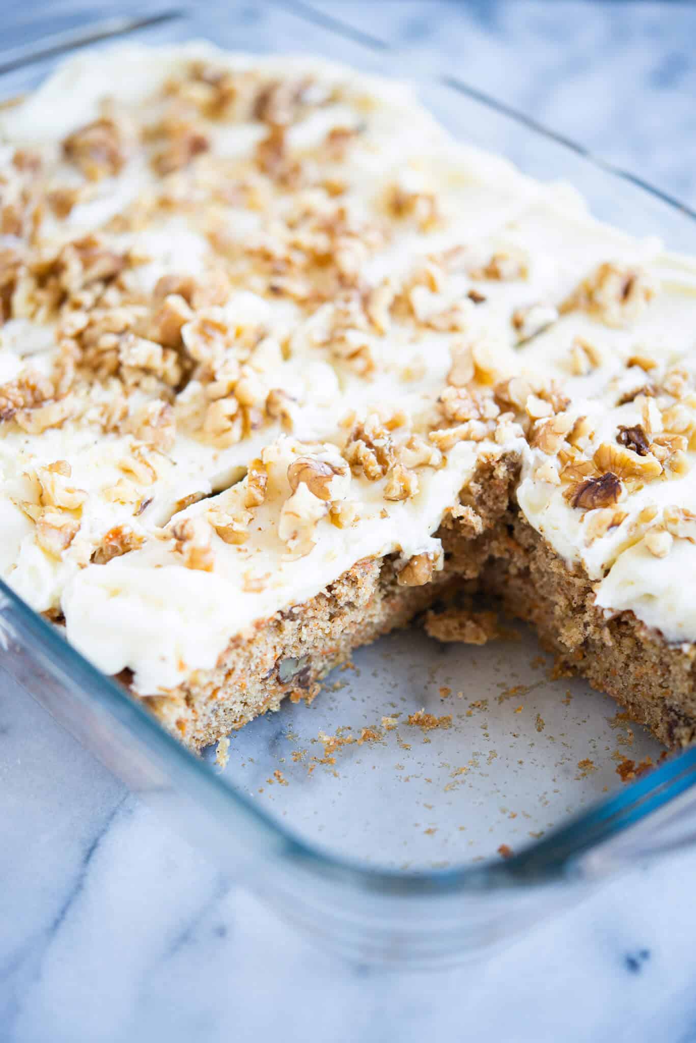 iced carrot cake cut into pieces with two pieces missing in a clear baking dish on a marble surface