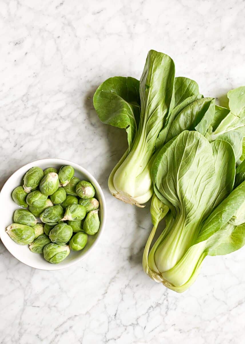 two stalks of raw bok choy laying next to a bowl of raw brussels sprouts on a marble surface