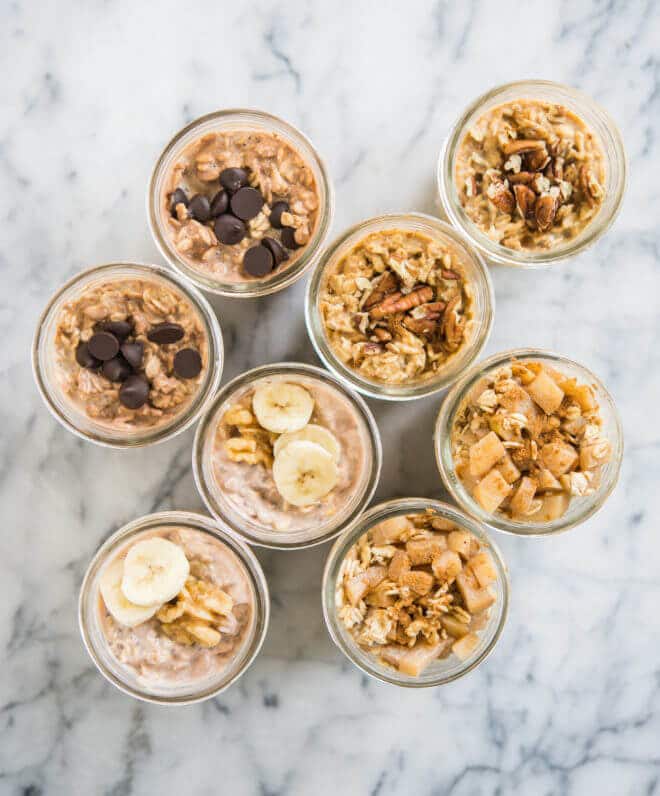 8 glass mason jars filled with overnight oats with various toppings like diced apples, sliced bananas, chocolate chips, and pecans on a marble surface