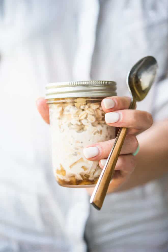 woman in a white shirt holding a mason jar full of overnight oats with a wooden-handled spoon