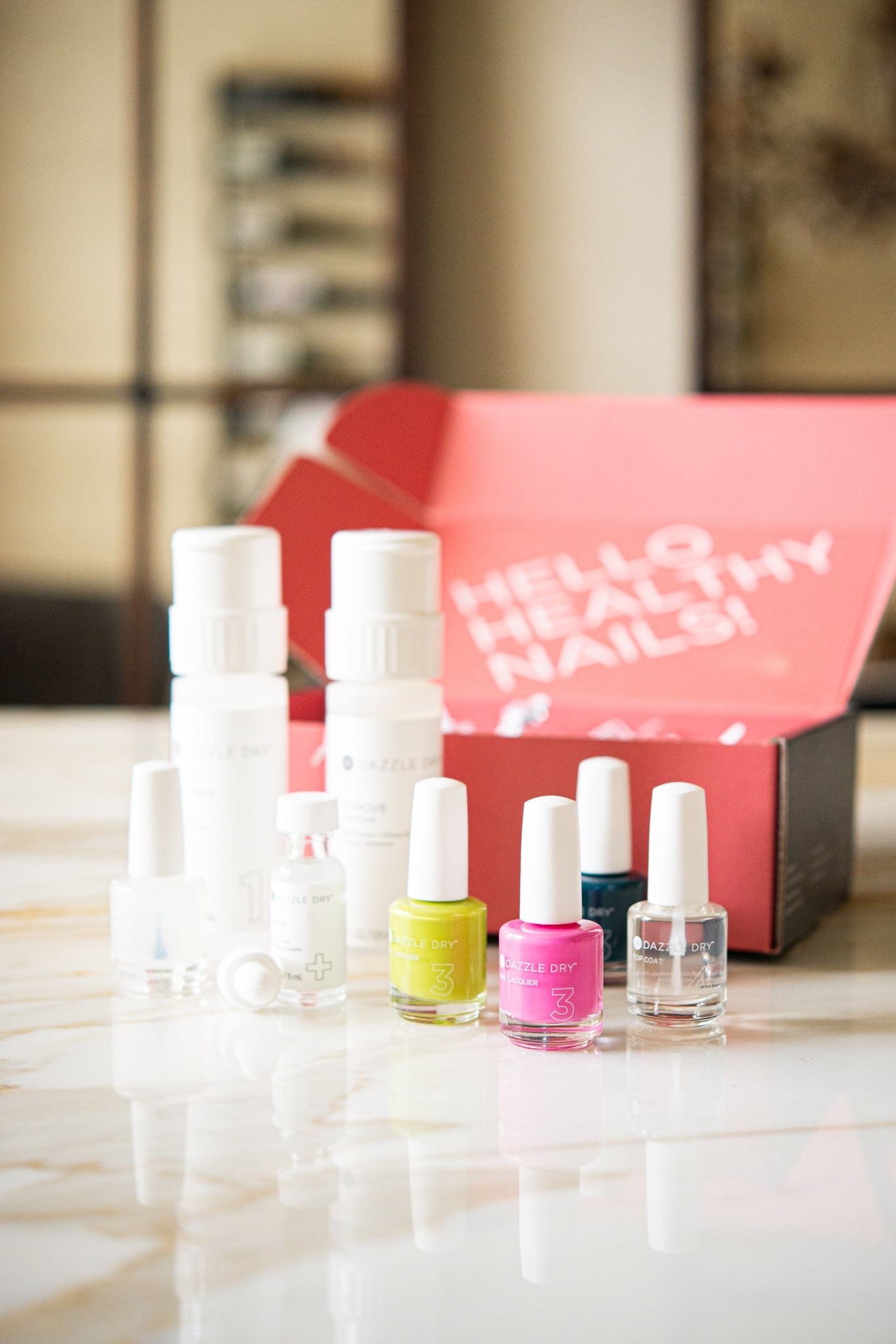 dazzle dry nail kit with three nail polishes, a top coat, and a remover, in front of a bright pink box